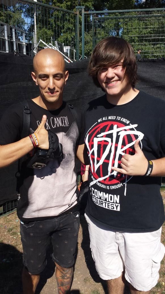 Two young men smiling and making the shaka hand gesture outdoors, with a fence and trees in the background. One has a shaved head, wearing a black T-shirt, and the other has long brown hair, wearing a black T-shirt with a graphic design.
