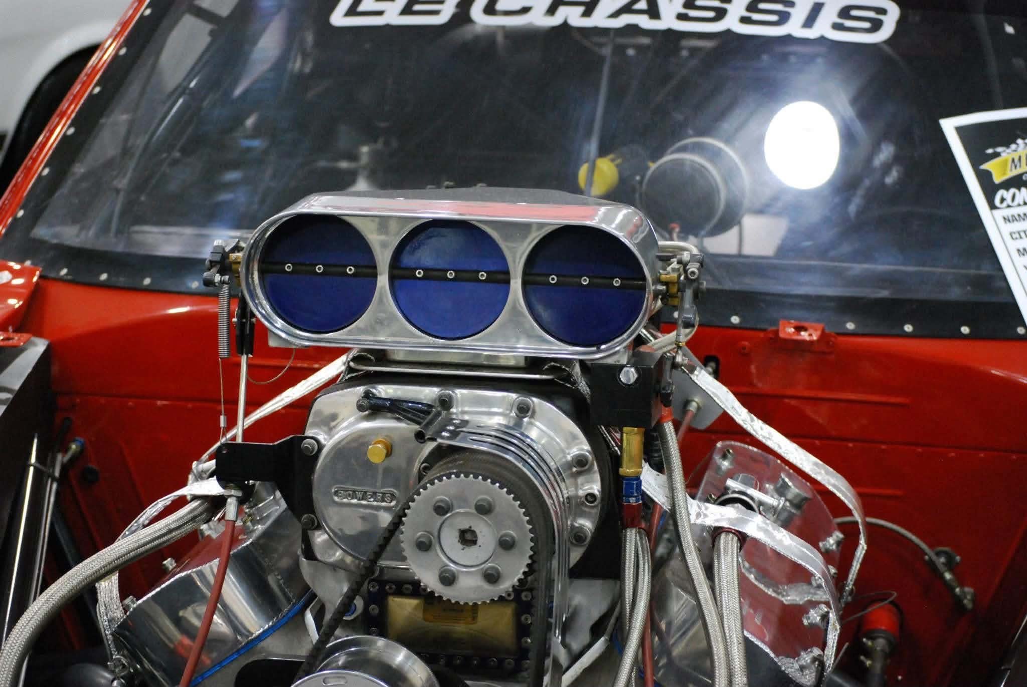 Close-up of a racing car engine, showing intake with three large circular openings, various metal components, and red bodywork in the background.