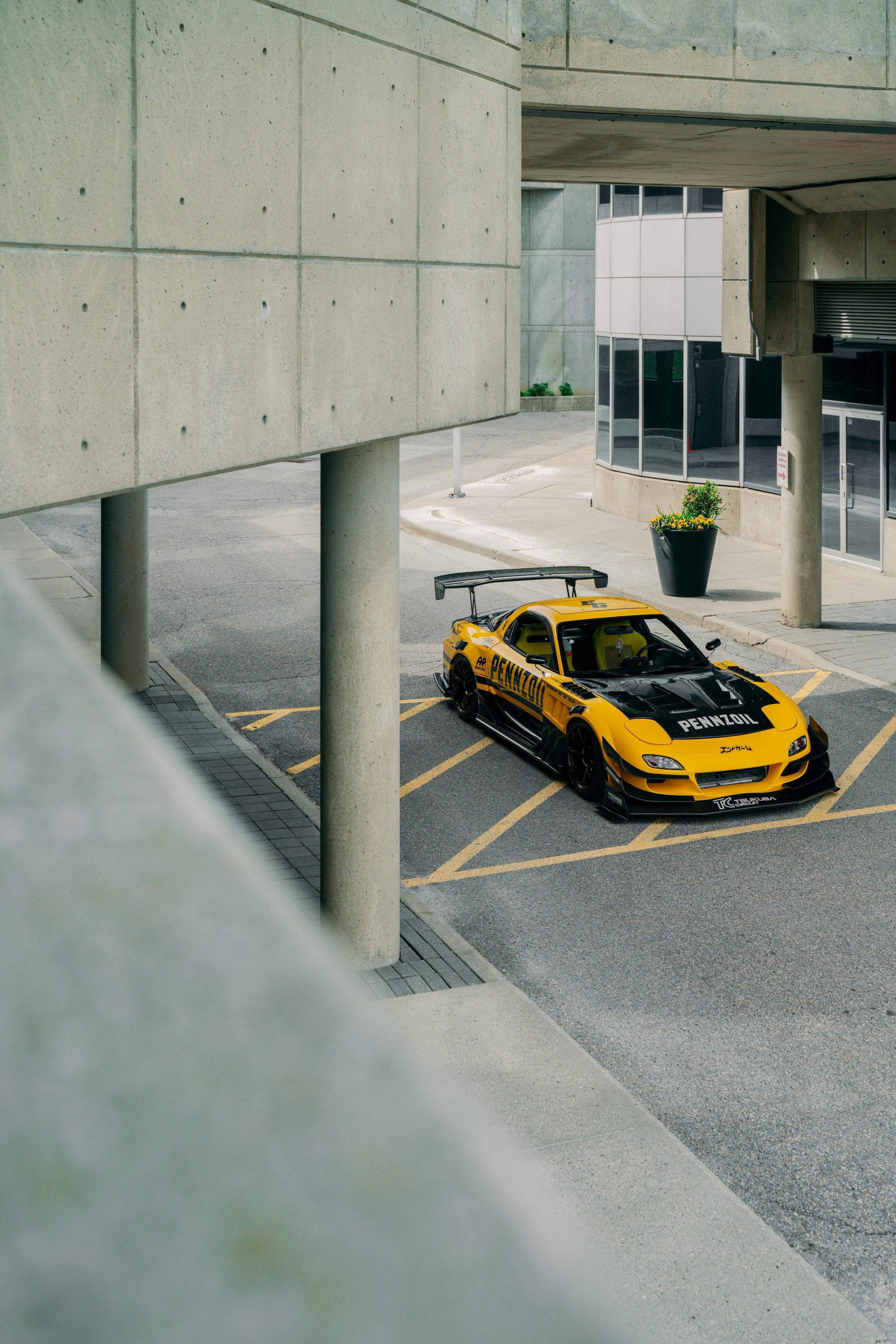 A yellow and black racing car with Pennzoil branding parked in a concrete parking lot near a building with large windows and a flower pot.