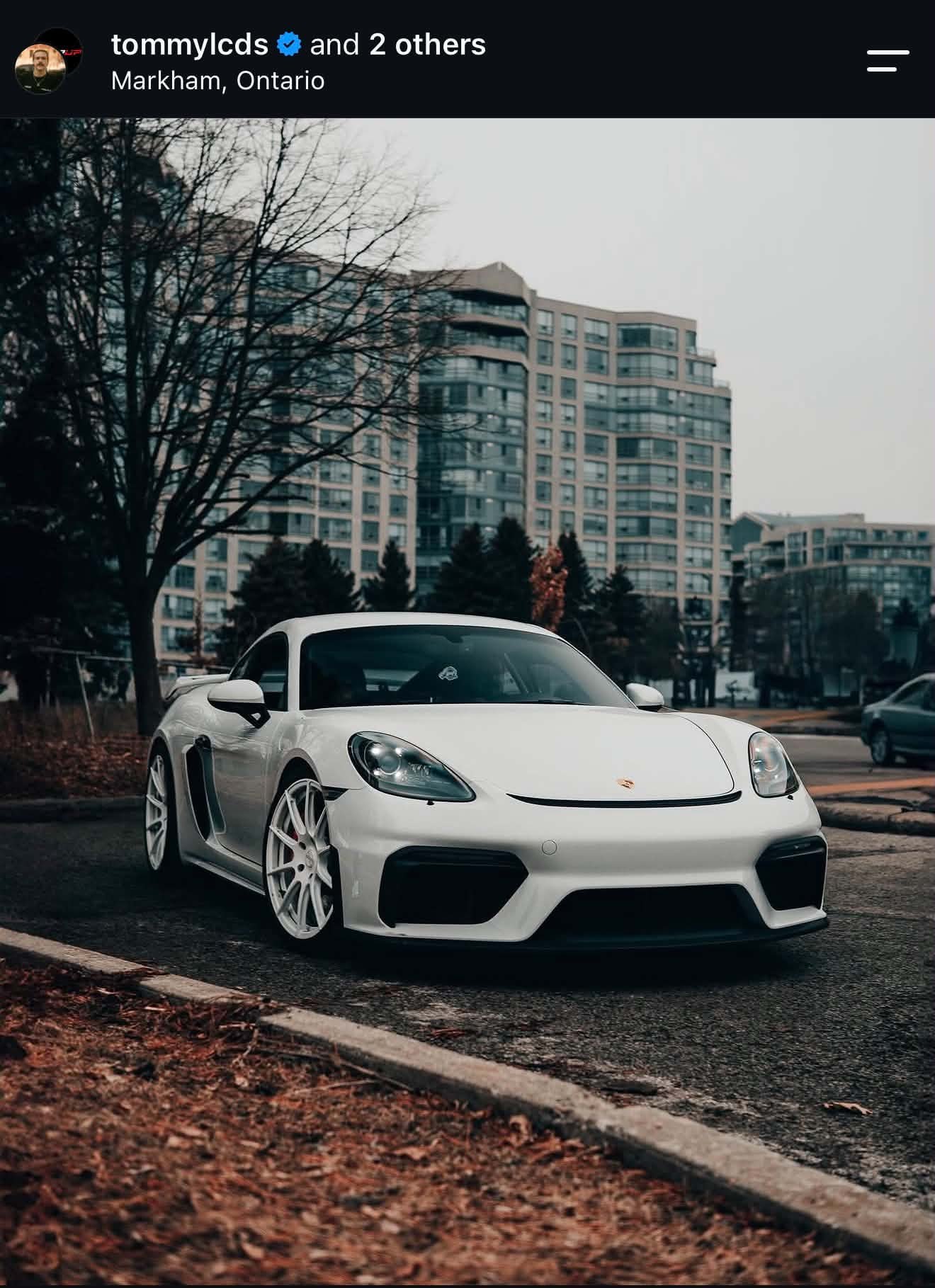 White sports car parked on street with tall residential buildings and trees in the background.