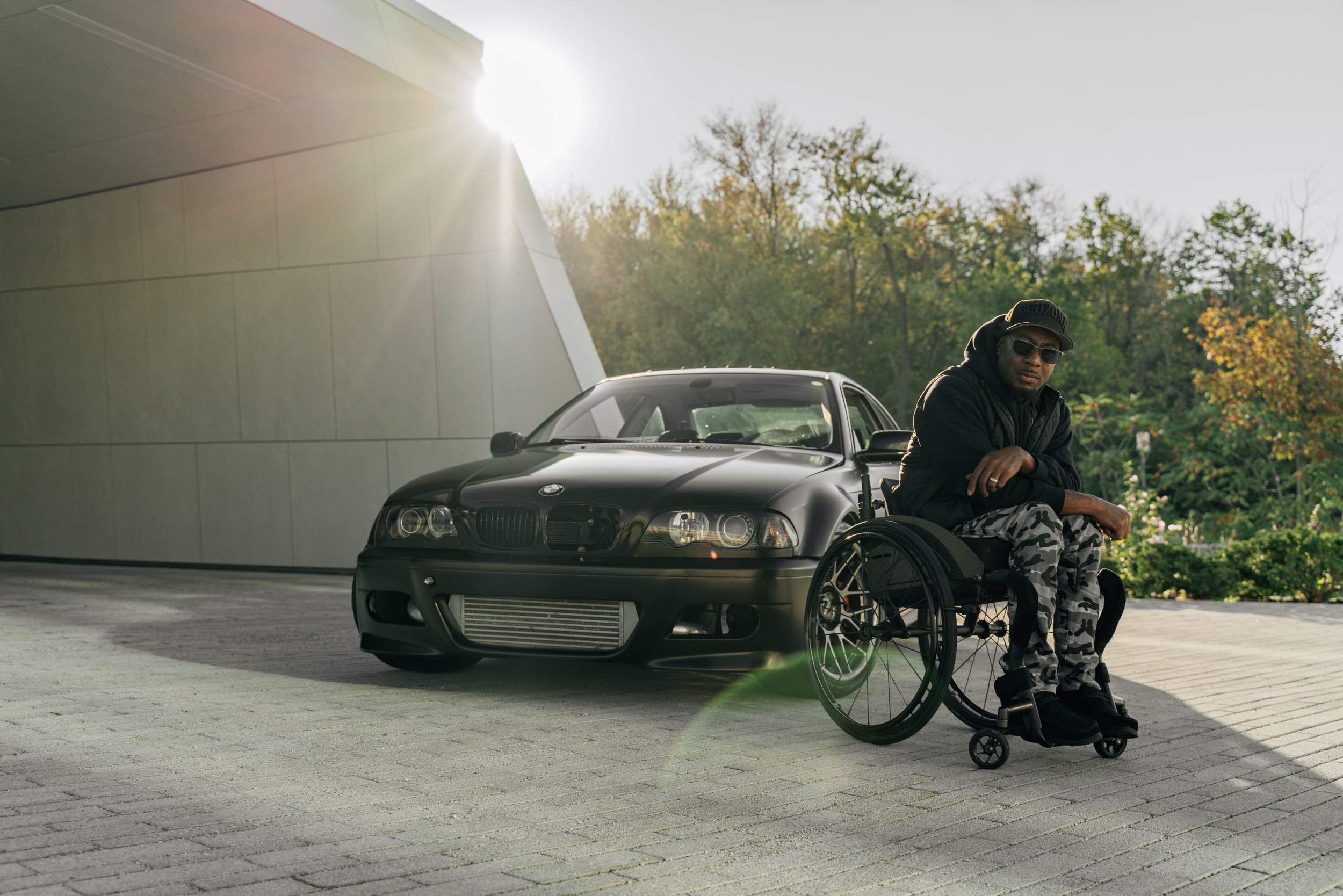 A man in a wheelchair with sunglasses and a cap sitting on a paved area in front of a black BMW car during daytime. Trees and a modern building with reflective surfaces are in the background, and sunlight creates a lens flare effect.