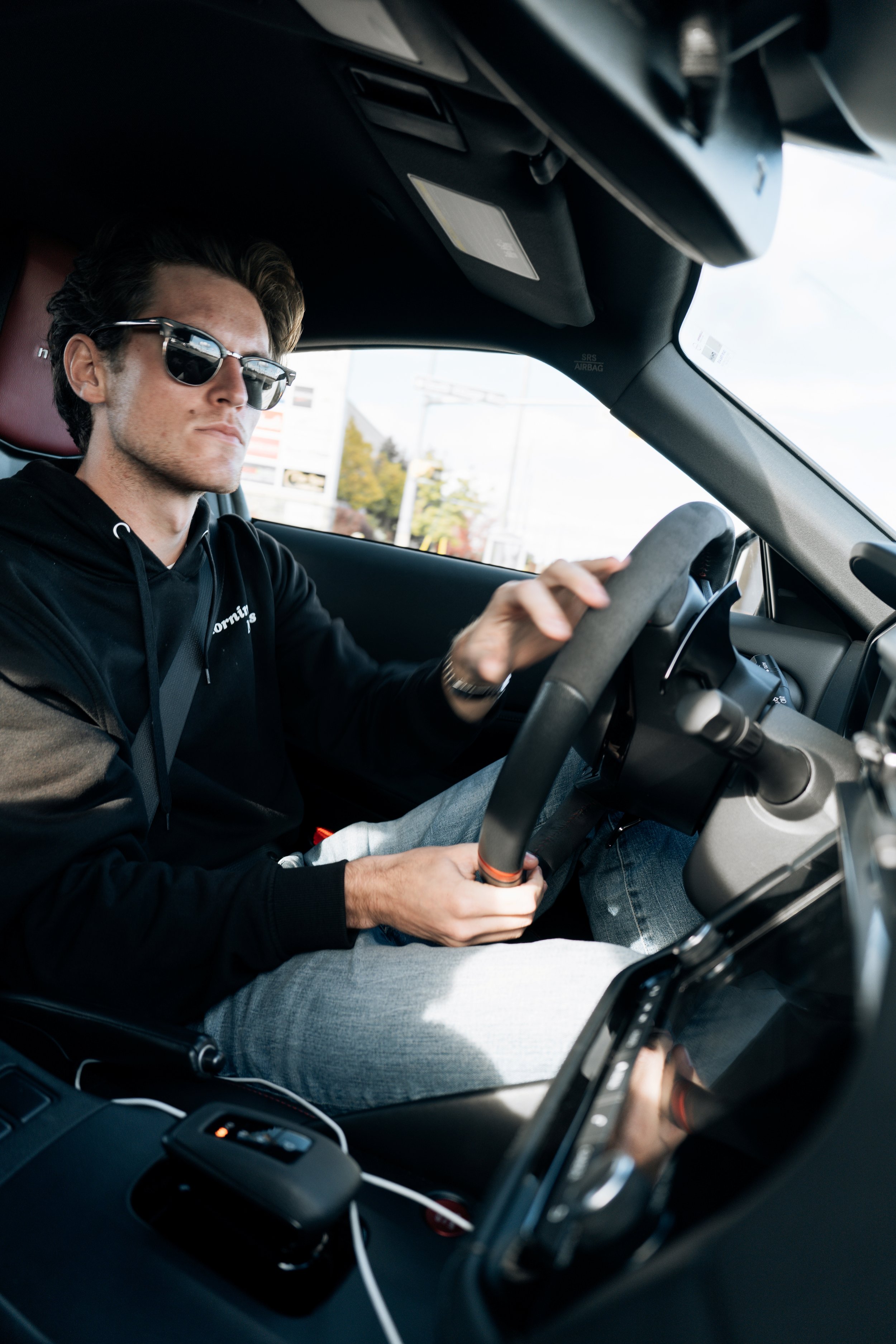 Young man with sunglasses sitting in car, holding steering wheel with one hand, wearing a black hoodie and grey pants, with a cityscape outside the window.