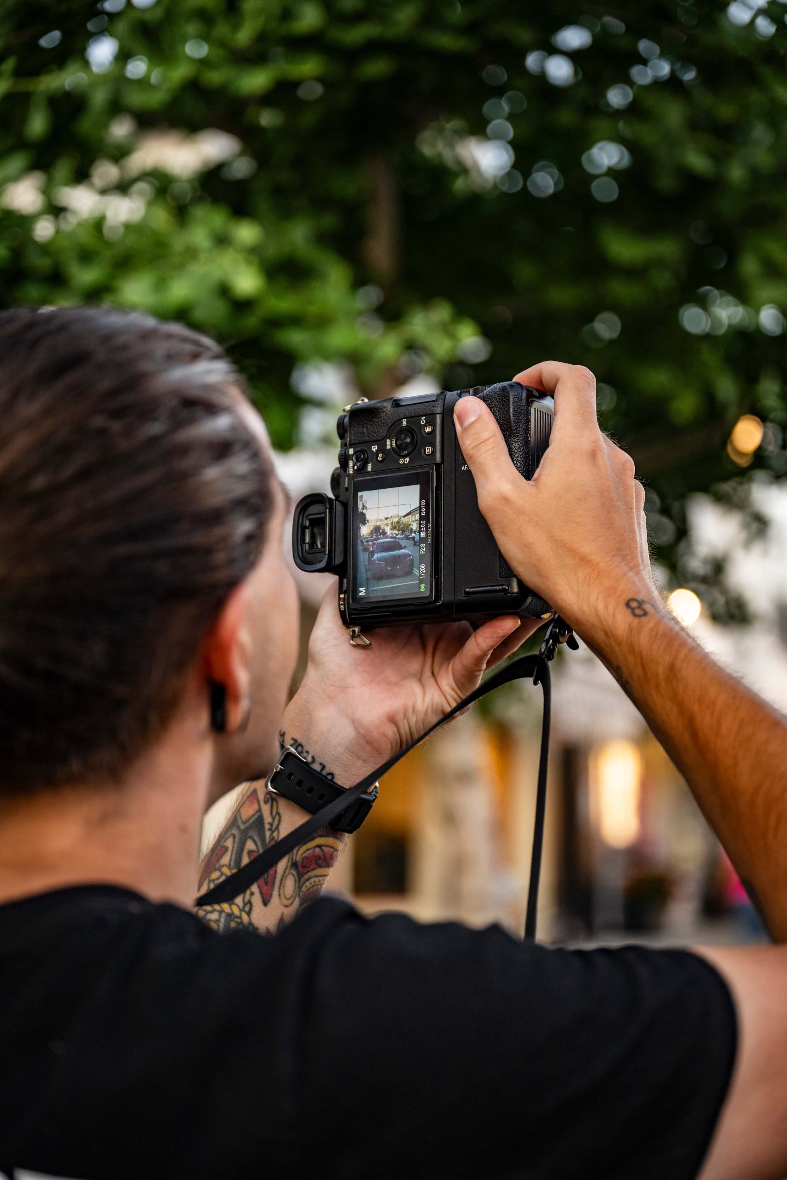 A person with brown hair holding a camera and taking a photo outdoors, with a blurred background of green trees and bokeh lights, wearing a black shirt, with visible tattoos on their arms.
