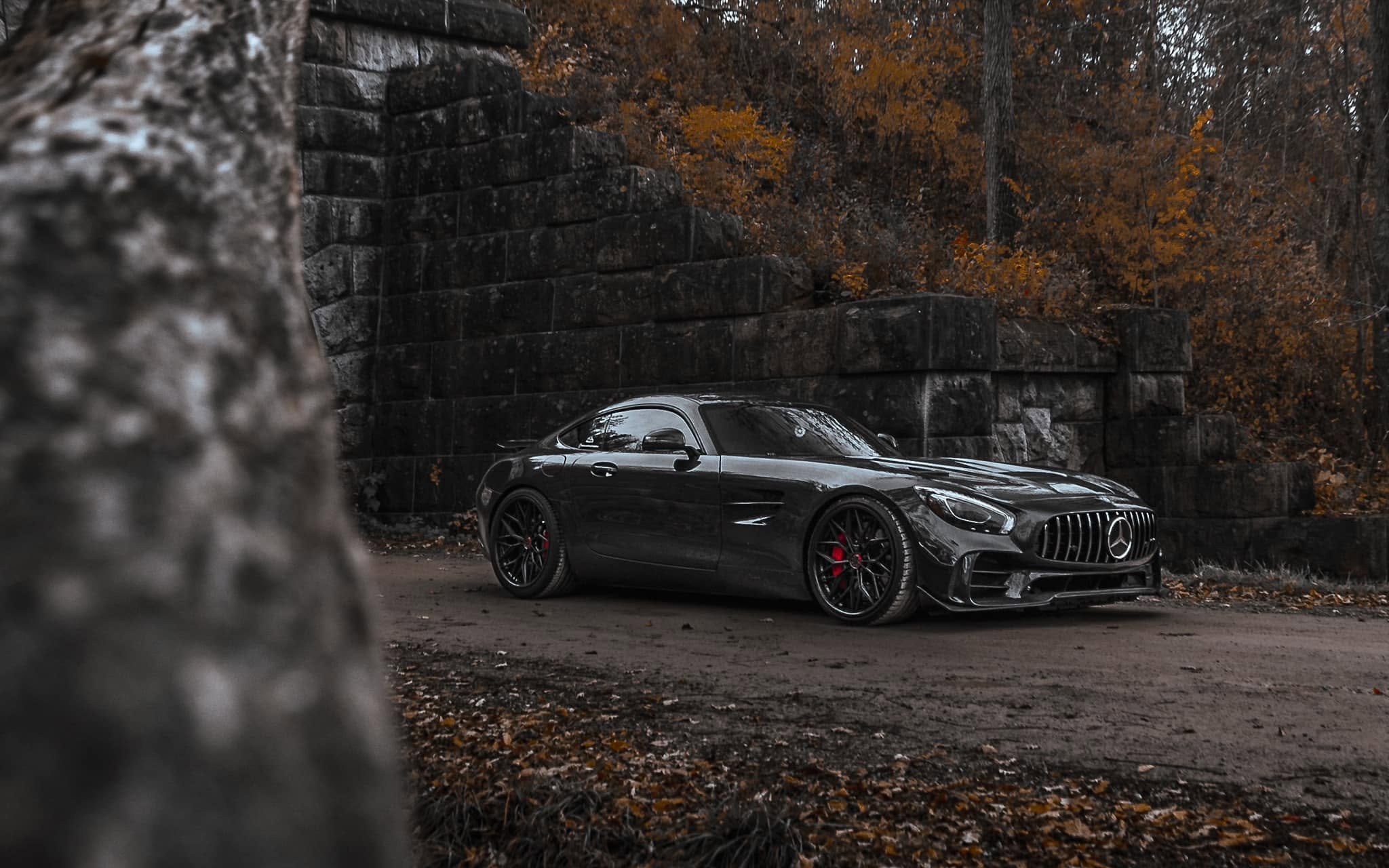 A black Mercedes-Benz sports car parked on a dirt area in front of a stone wall with fall foliage in the background.