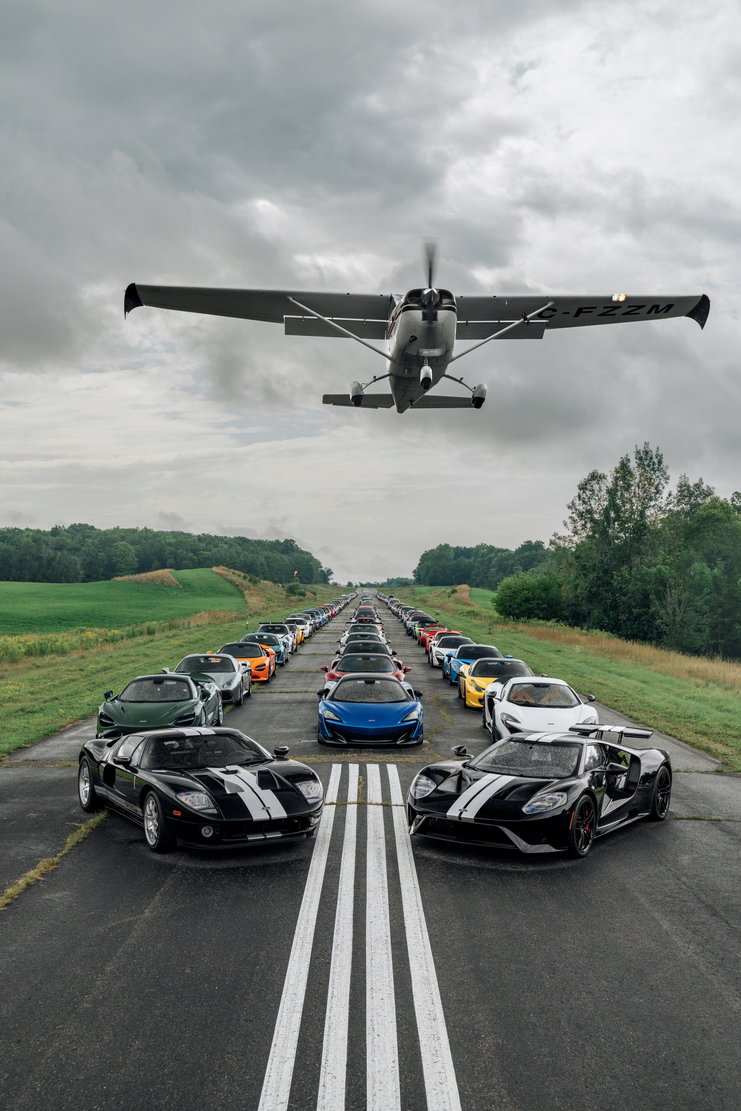 A collection of sports cars lined up in a parking lot, with two black cars featuring white racing stripes in the foreground, and a small airplane flying low overhead against a cloudy sky.