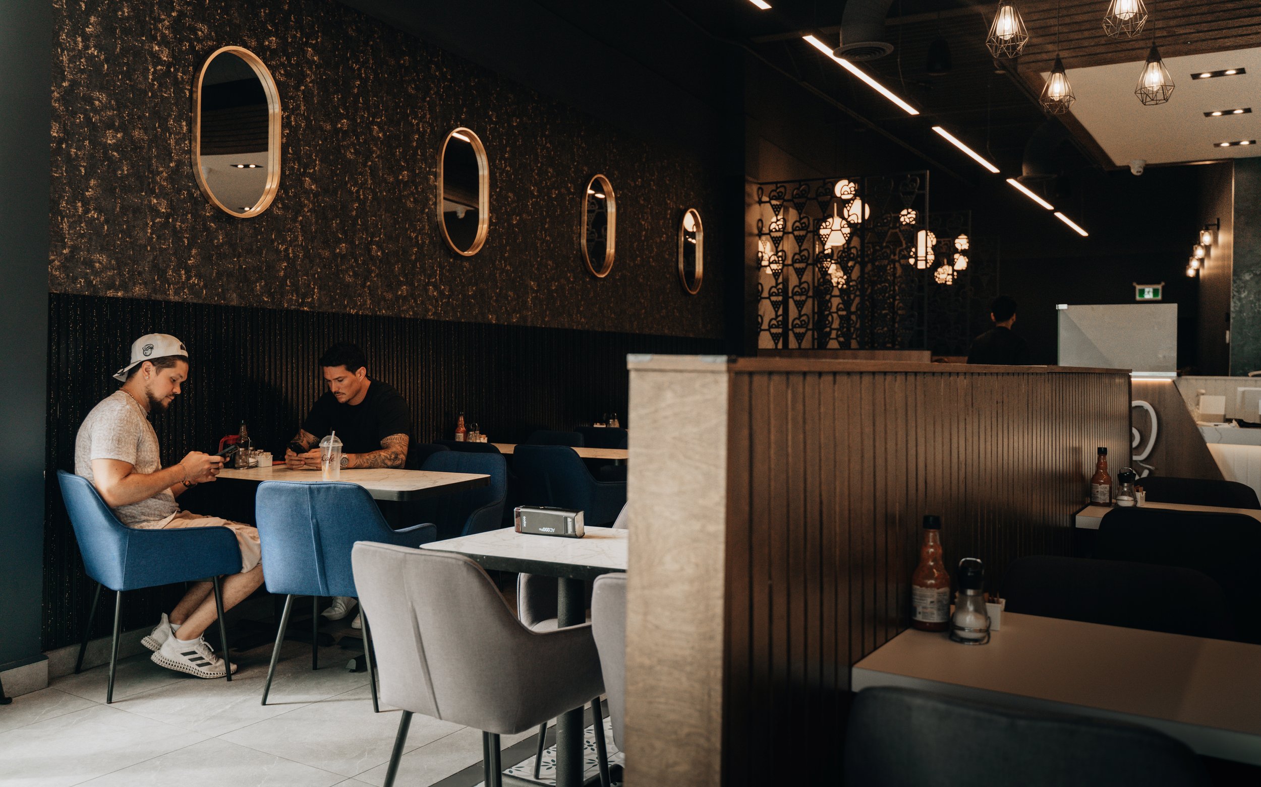 A modern, dimly lit restaurant with dark walls, circular mirrors, and decorative lighting fixtures. Two men are seated at a table, looking at their phones, with condiments on the table.