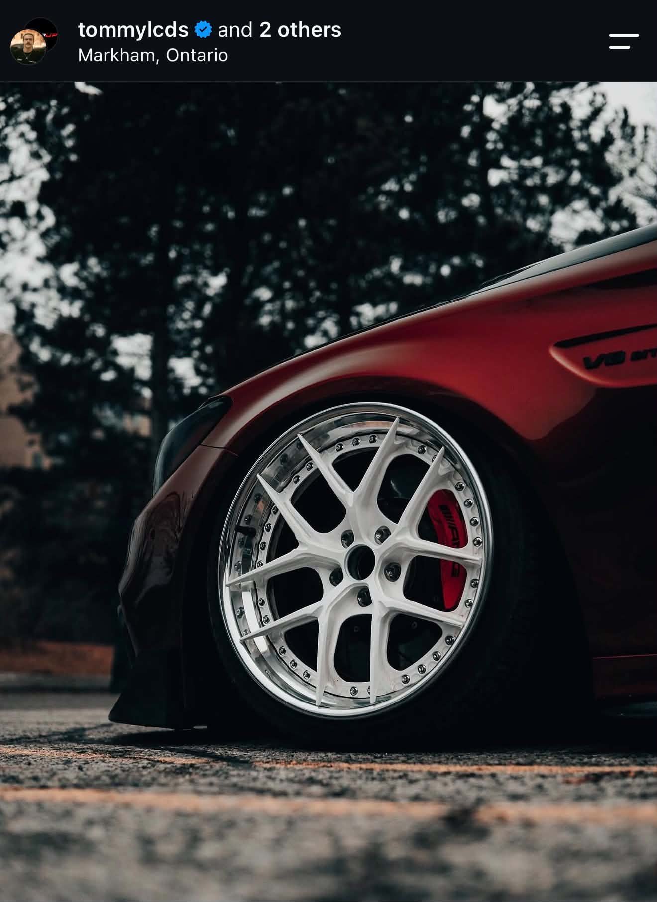 Close-up of a red sports car's front wheel with white rims and red brake caliper, parked on a textured road with trees in the background at dusk.