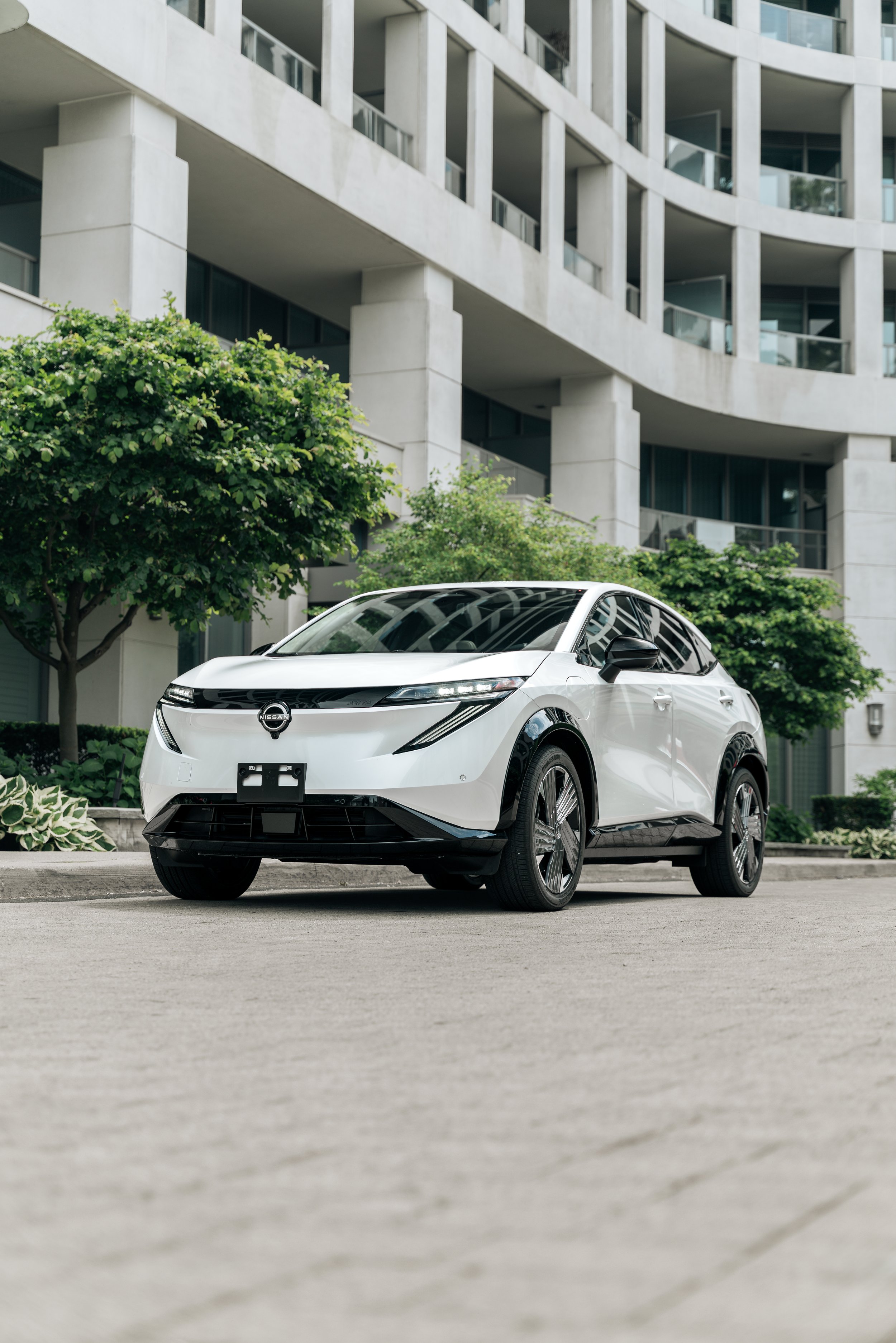 A white Nissan electric vehicle parked in front of modern residential buildings with green trees and shrubs nearby.