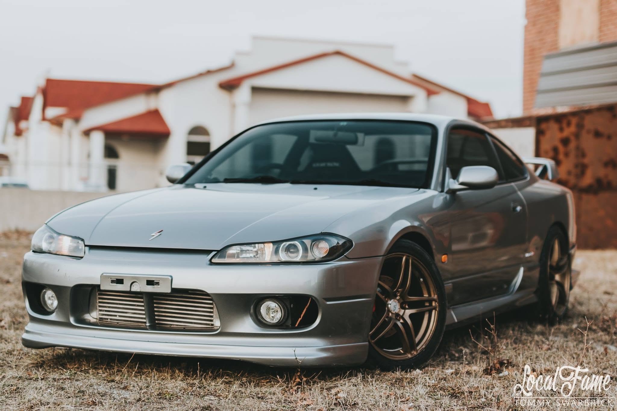 A silver sports car parked on a grassy area with houses and a cloudy sky in the background.