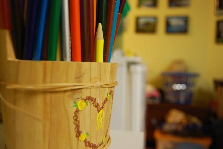 A wooden container with a heart-shaped decoration painted on it, filled with colored pencils, sitting on a surface in a room with yellow walls and framed pictures.