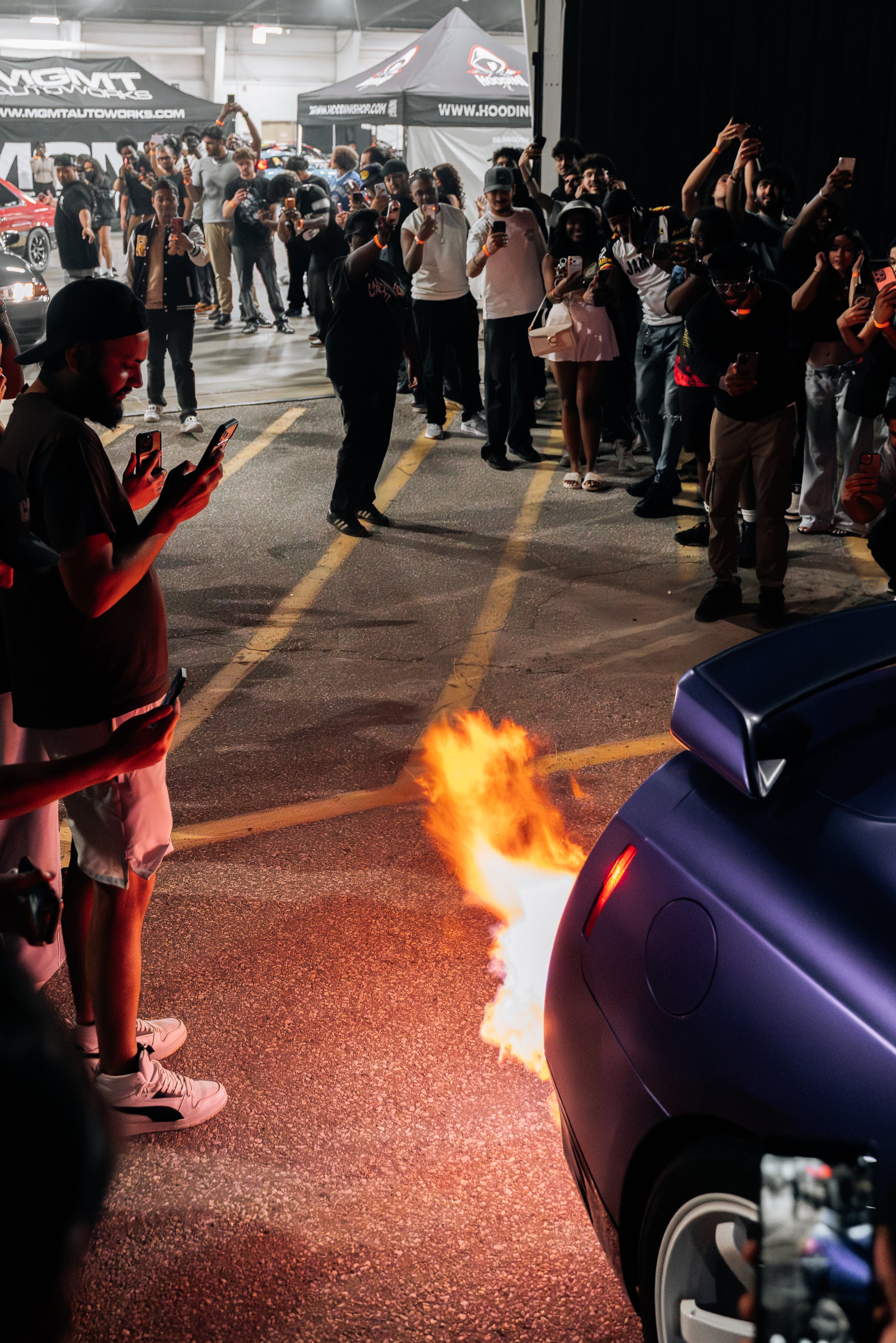 A crowd of people inside a garage or event space, many taking photos or videos with their phones, watching a small fire near a purple car.