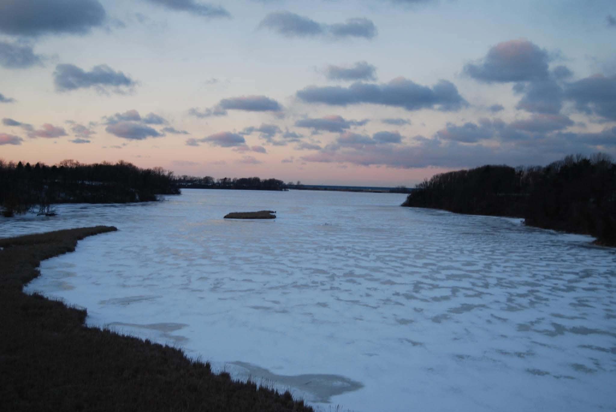 A river with snow-covered banks and small islands, under a cloudy sky during dusk or dawn.