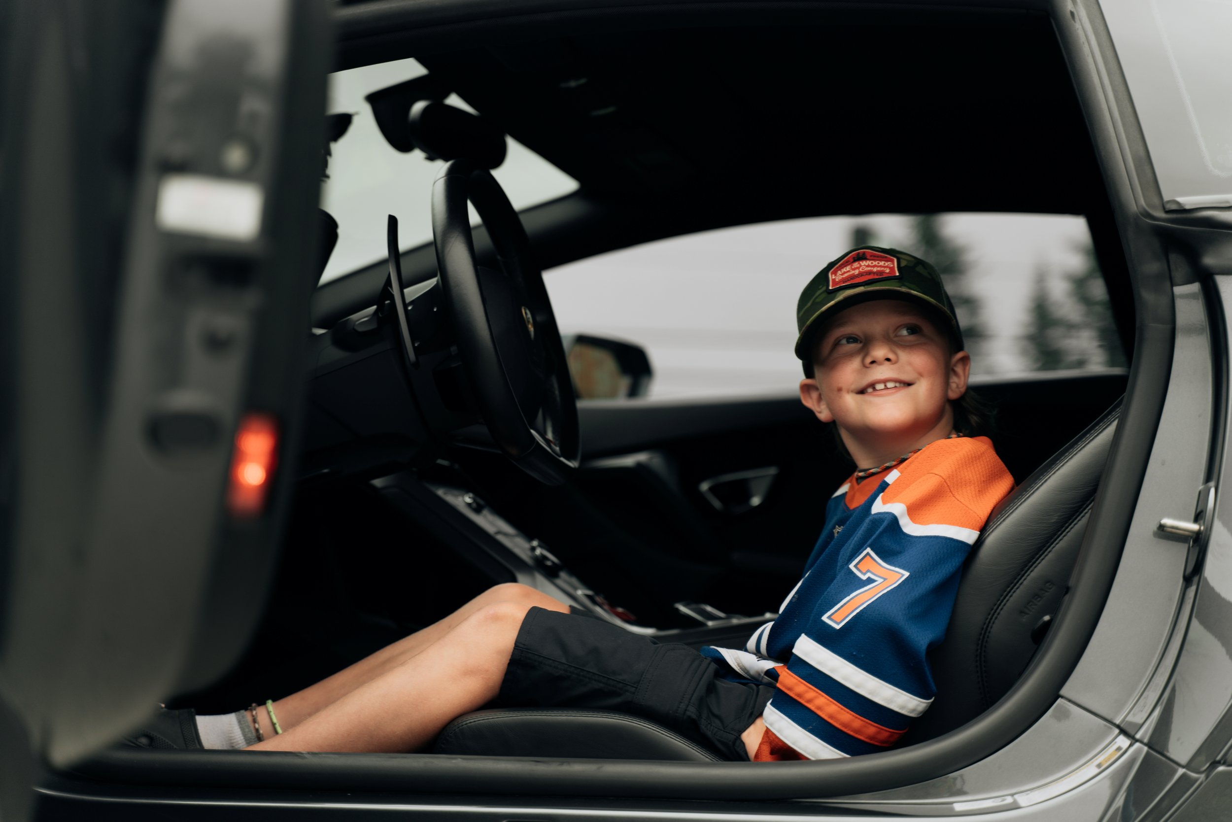 Young boy smiling while sitting in the driver's seat of a vehicle, wearing a cap and sports jersey, with a person reaching over to adjust something inside the car.