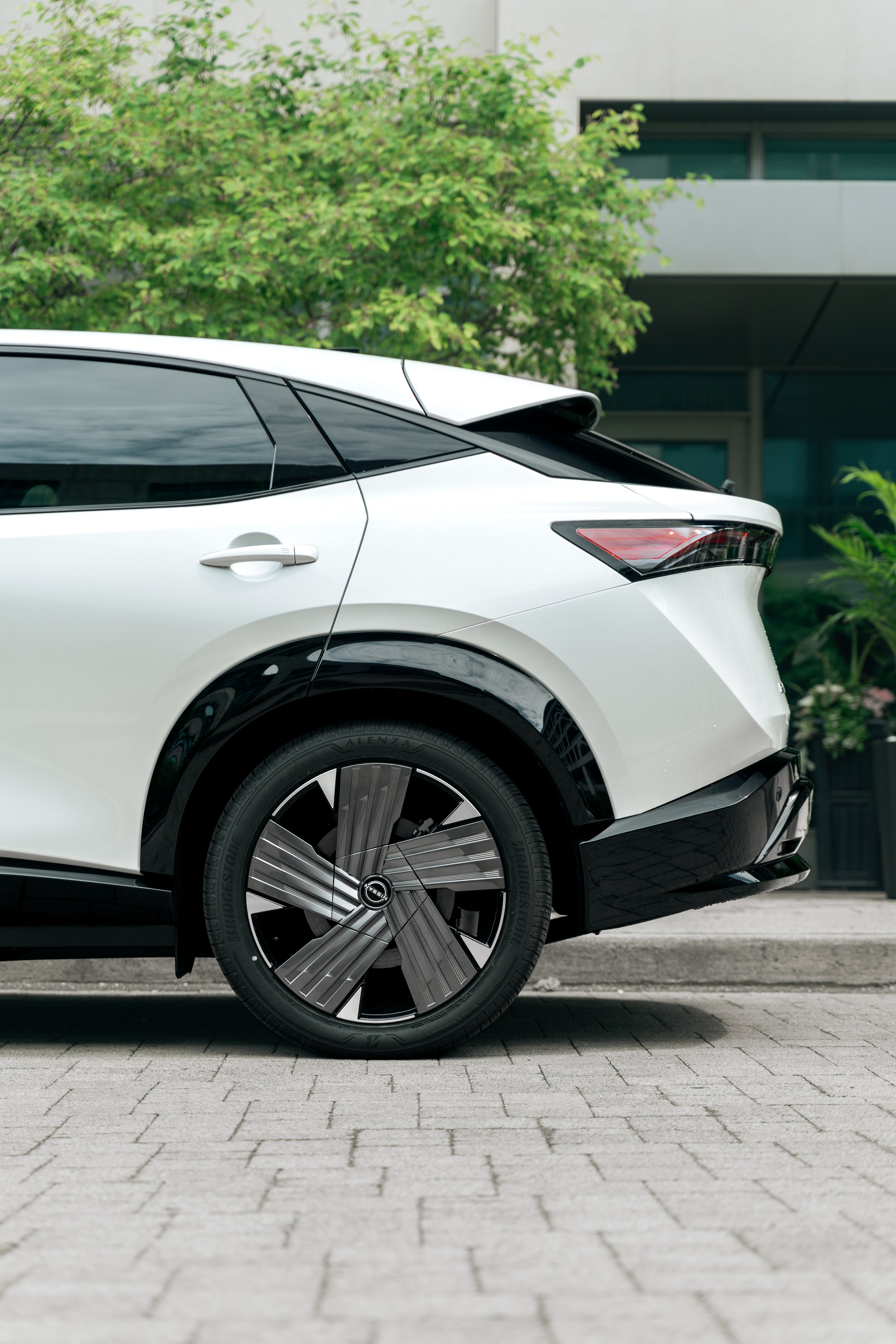 Close-up of a modern white electric vehicle parked on a paved street, with a decorative wheel cover, black trim, and a background featuring green trees and a building facade.