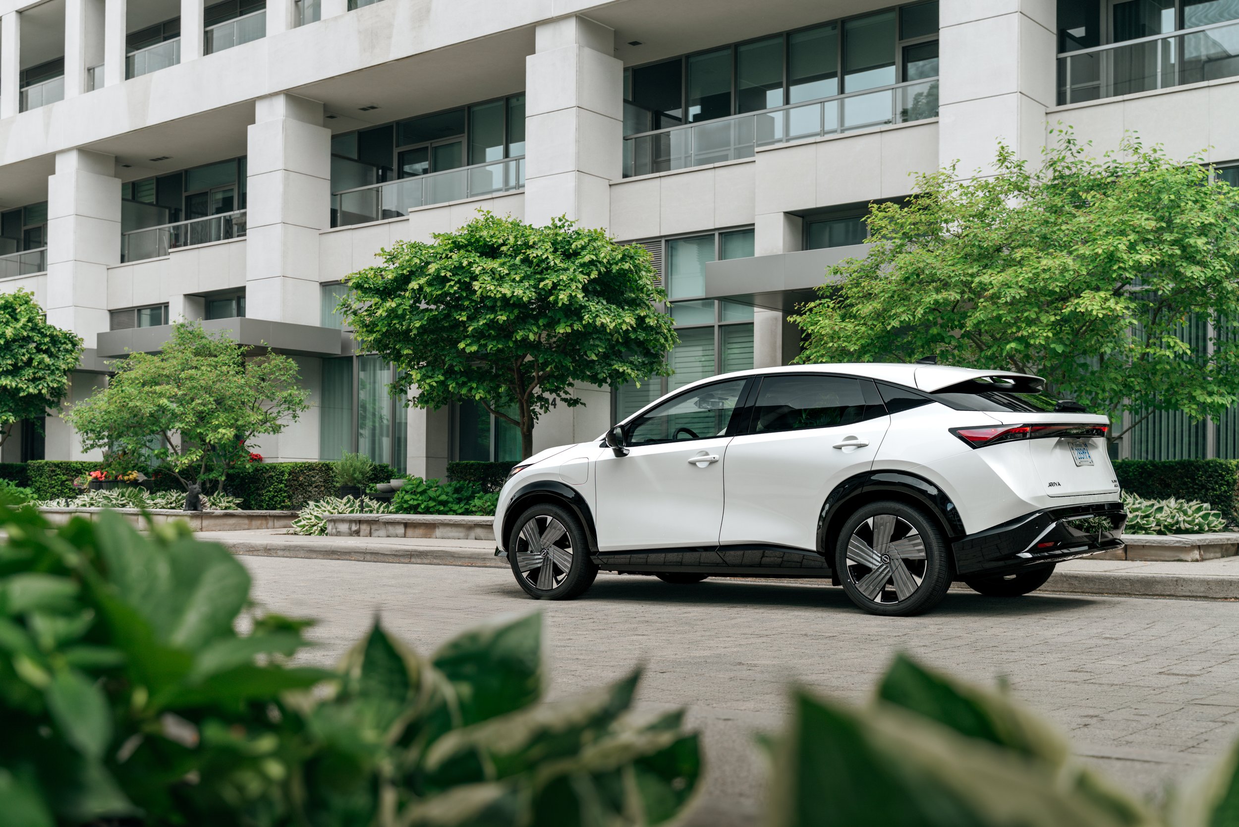 A white electric vehicle parked in front of a modern apartment building with balconies and large windows, surrounded by trees and greenery.