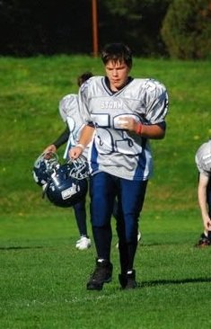 Young football player in uniform walking on a grassy field carrying a helmet and football.