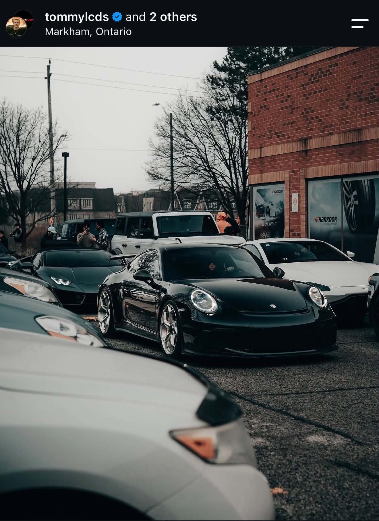Black and white Porsche sports car parked on the street with other cars nearby, storefront with advertisements, leafless trees, and a brick building, in Markham, Ontario.
