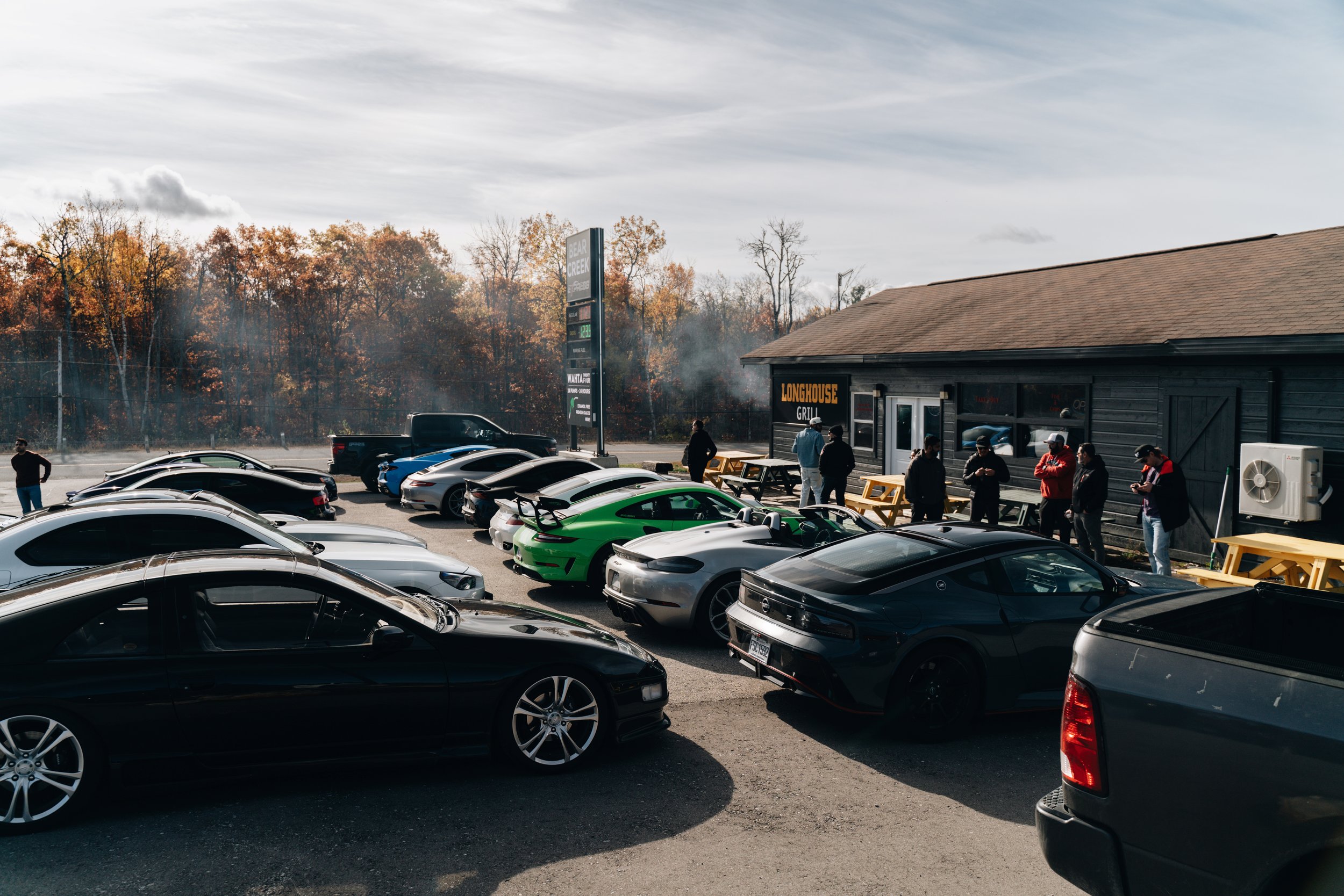 A parking lot filled with various cars, with some people standing near a black building that has a sign saying 'Longhouse Grill'. In the background, there is a sign for Bear Creek and trees with fall foliage under a cloudy sky.