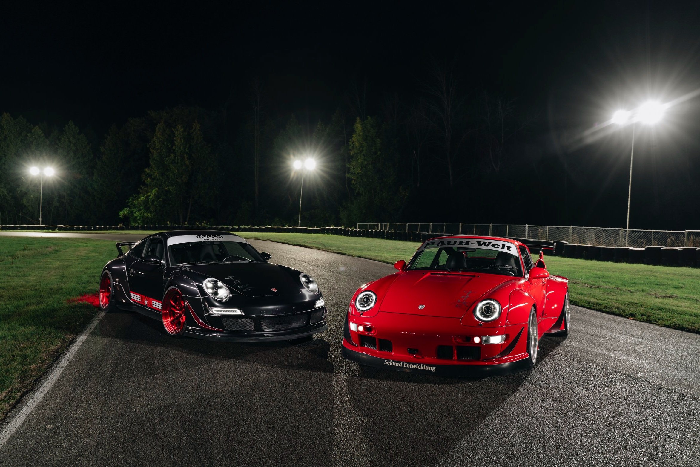 Two sports cars, a black one with red accents and a red one, are parked on a race track at night under bright stadium lights, with trees in the background.
