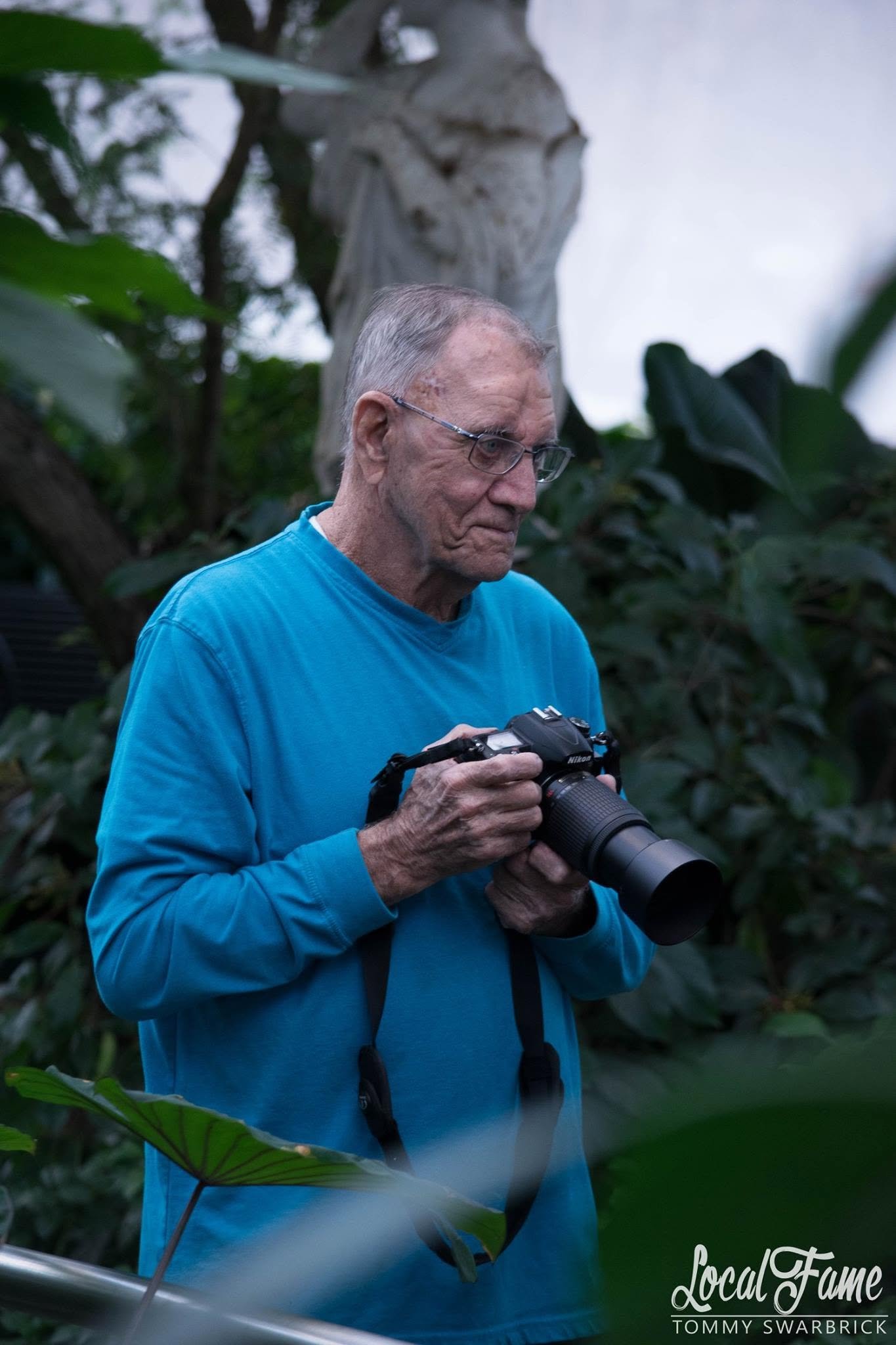 An elderly man with gray hair and glasses, wearing a blue long-sleeve shirt, looks down at a camera he is holding in his hands, surrounded by lush green foliage.