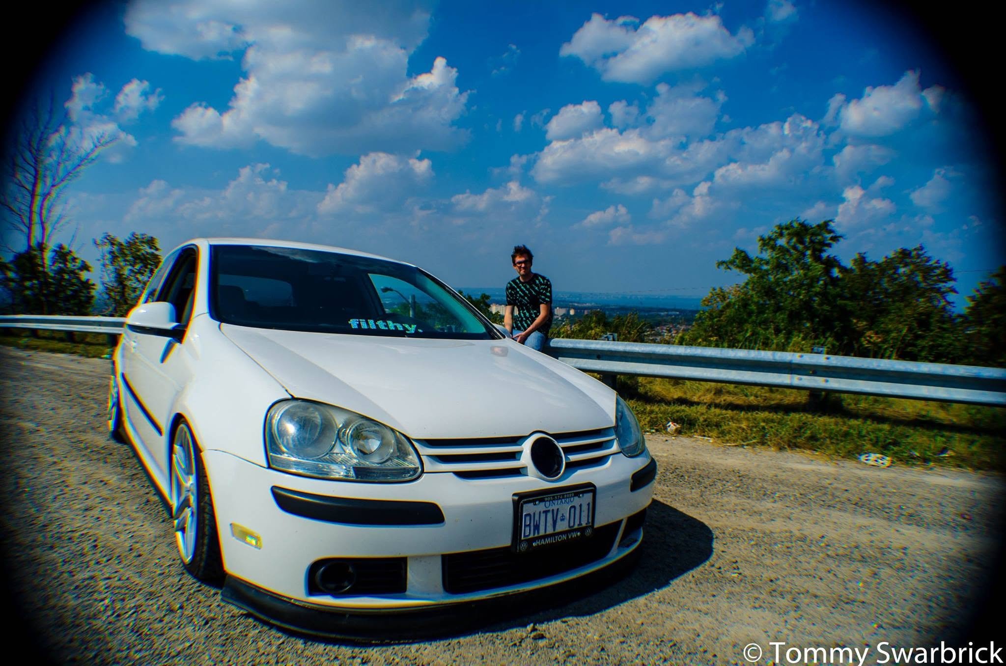A white car with a person sitting on the rear of the car, overlooking a scenic view of trees and a city in the background, under a partly cloudy sky.
