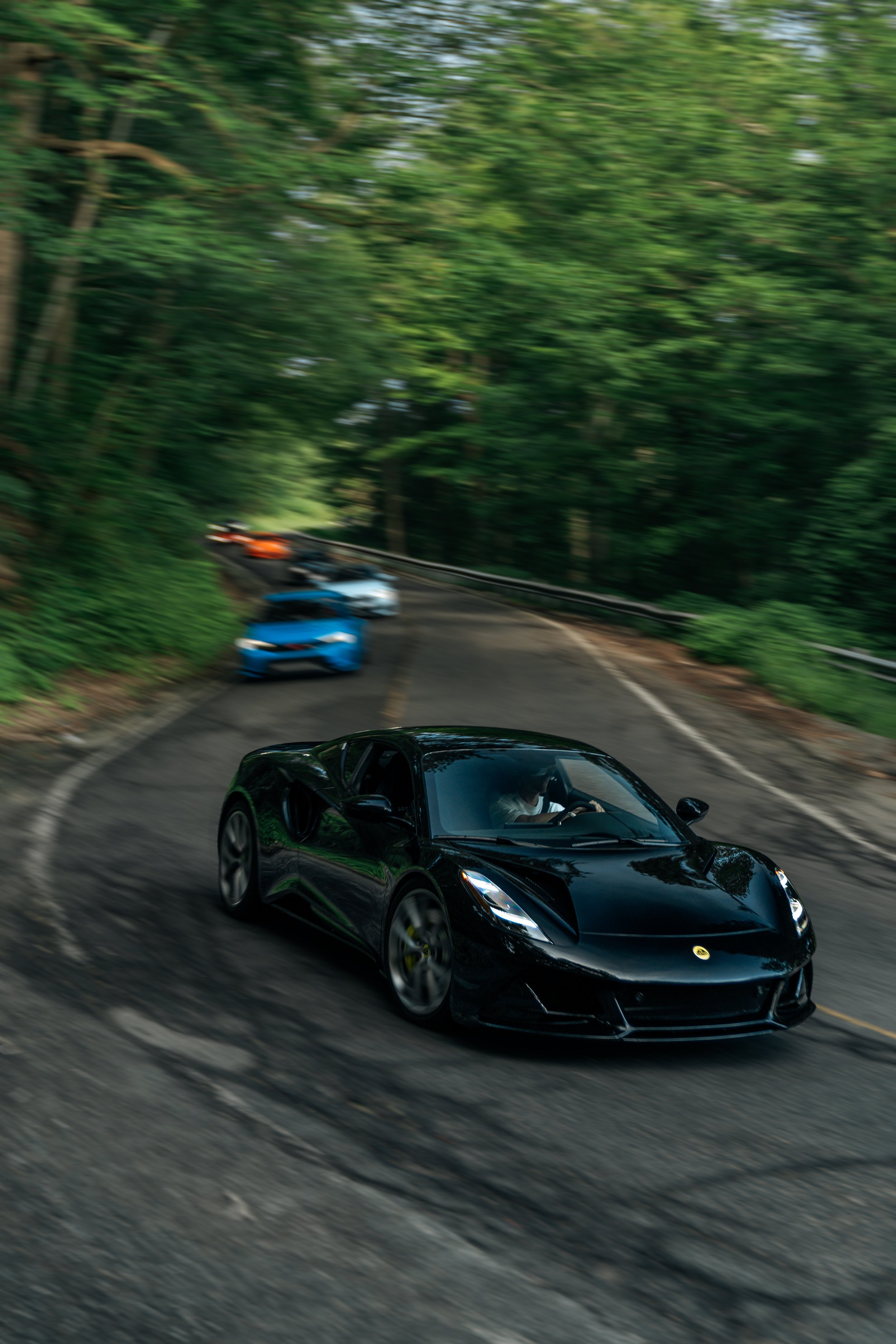 A sleek black sports car driving on a winding forest road surrounded by green trees, with other colorful sports cars following behind, motion blur indicating speed.