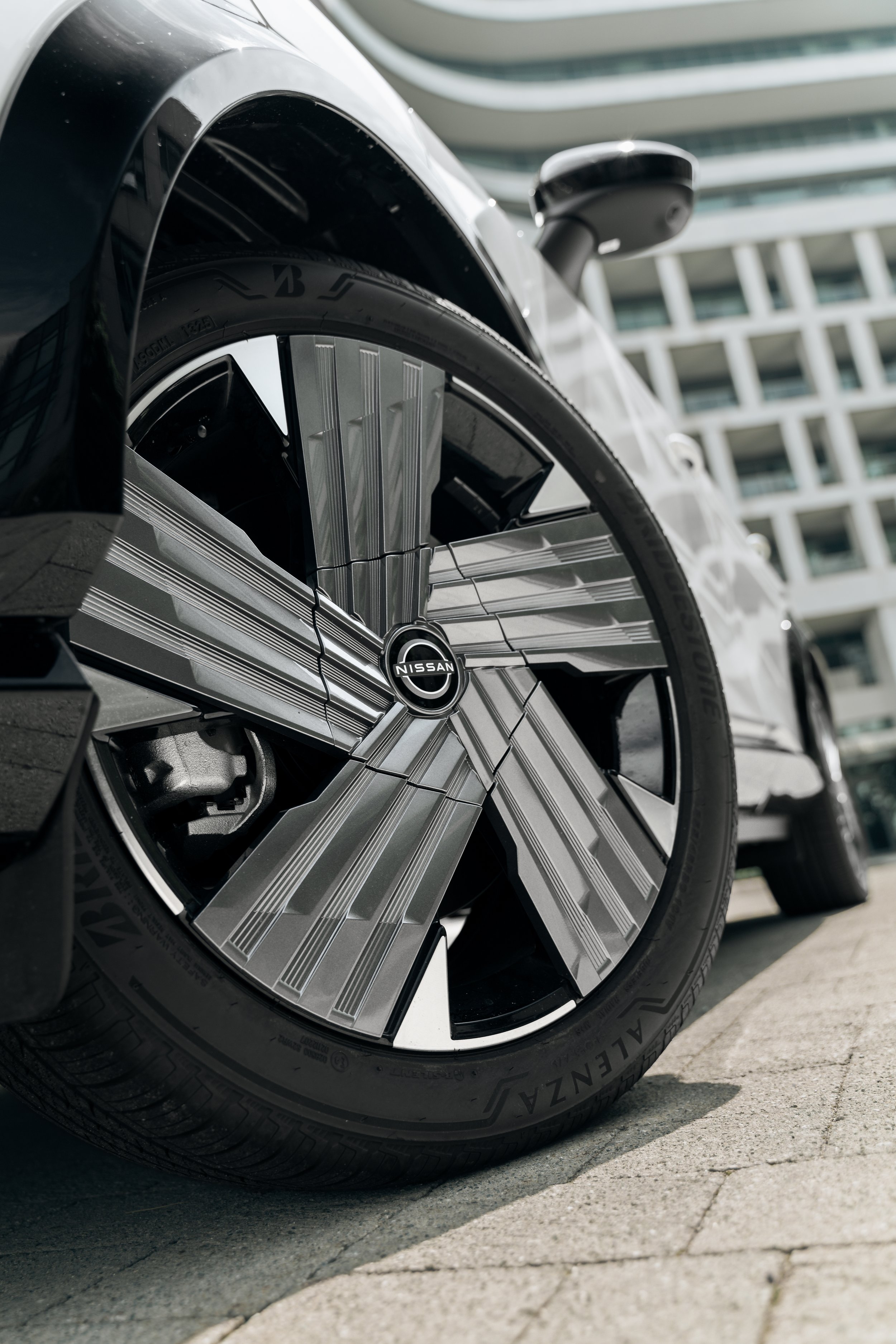 Close-up of a car's front tire and wheel with Nissan logo, parked on a paved surface, with modern building in background.