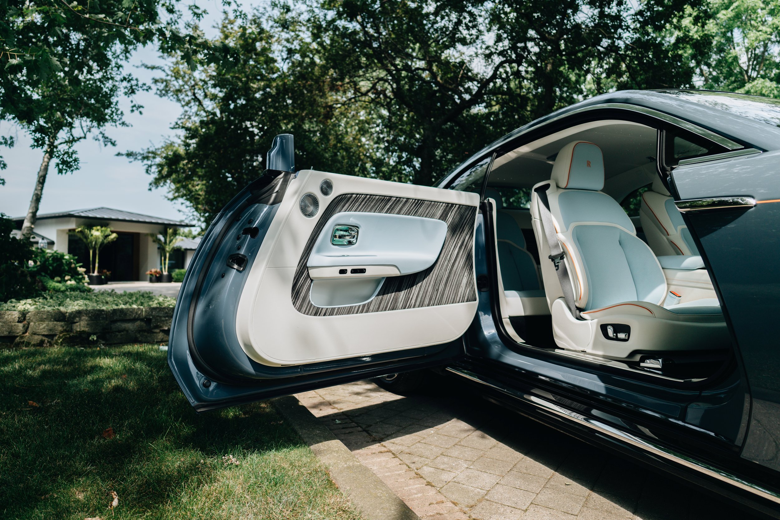 Interior of a luxury car with open door, showing white leather seats and wood trim, parked outside a house with a front yard and trees.