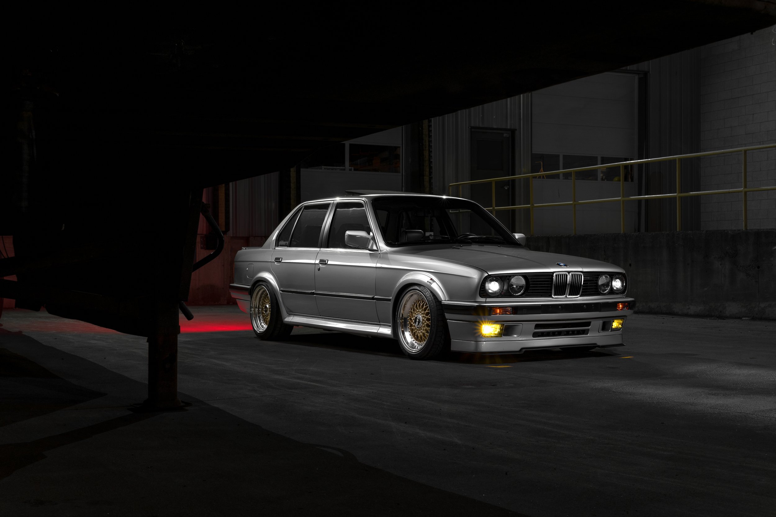 A silver classic BMW car with gold wheels parked in a dark, industrial-looking garage, illuminated by lights from the front of the vehicle.