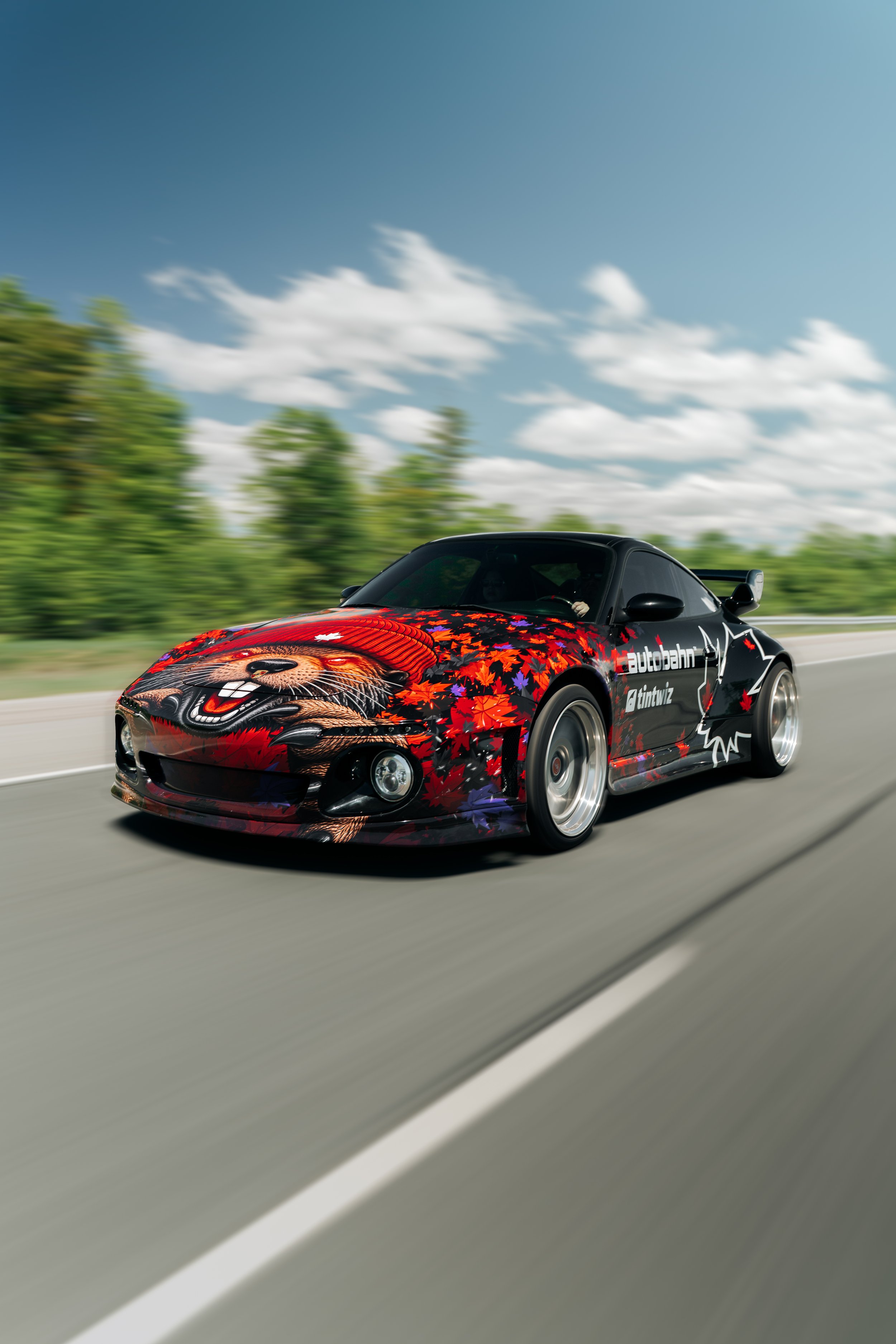 A black sports car with a colorful, autumn-themed design featuring a bear face and maple leaves, racing on a highway with motion blur behind it, under a partly cloudy sky.
