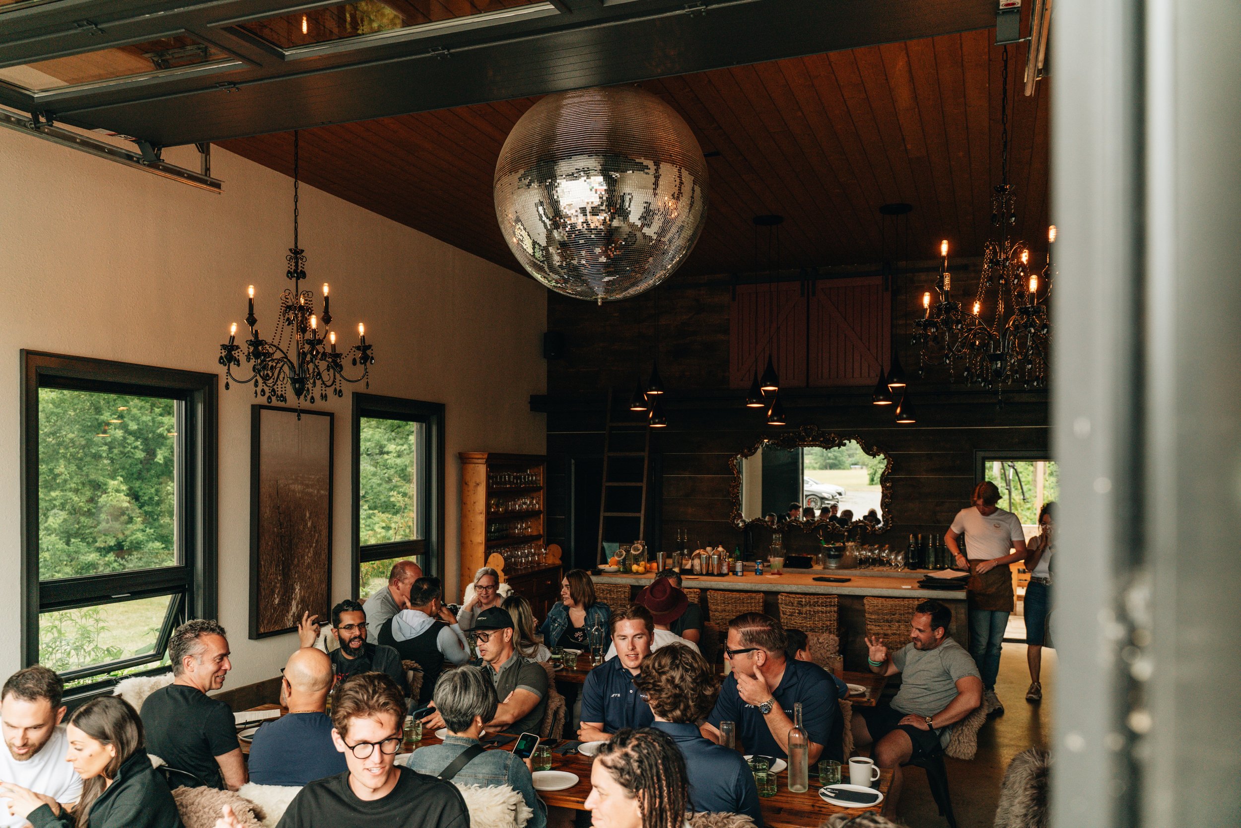 Interior of a lively restaurant or cafe filled with seated people enjoying their meals and conversations. The room features large windows revealing greenery outside, a wooden ceiling, elegant chandeliers, and a large decorative disco ball hanging from the ceiling.