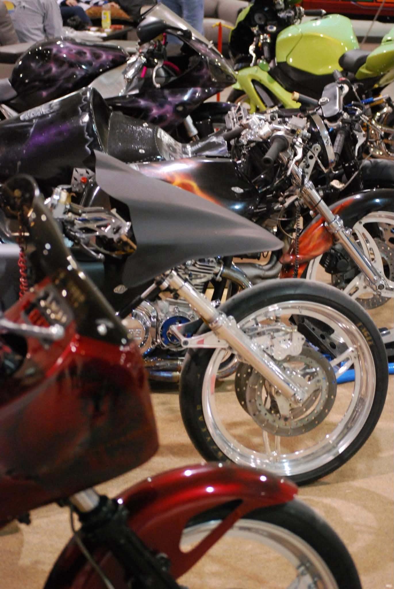 Multiple motorcycles parked indoors, including a black bike with a purple and pink flame paint job, a yellow bike, and a red motorcycle, with a blurred background of people and tables.