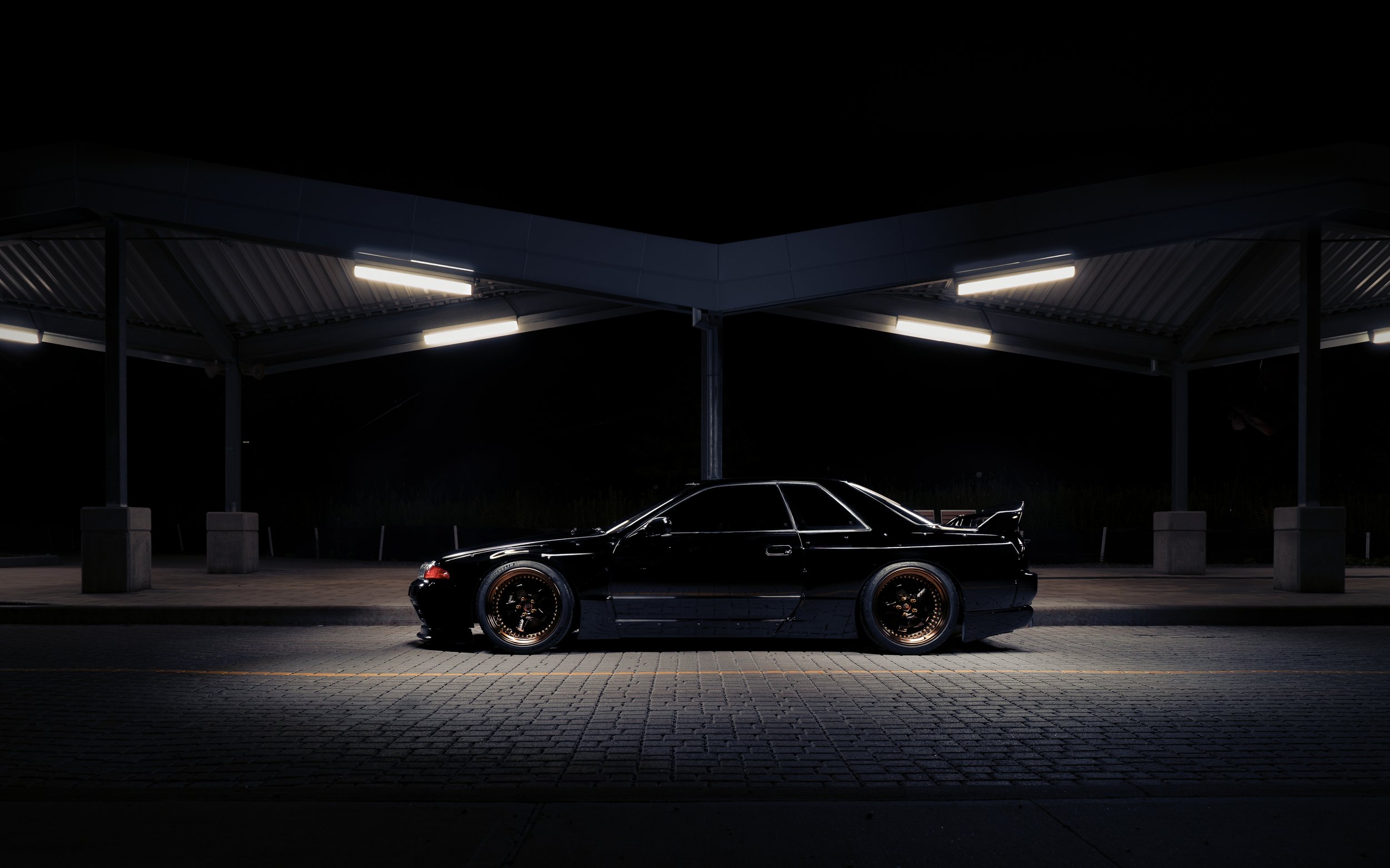 A black sports car parked at night under a metal-roofed canopy with bright lights, dark sky, and paved ground.