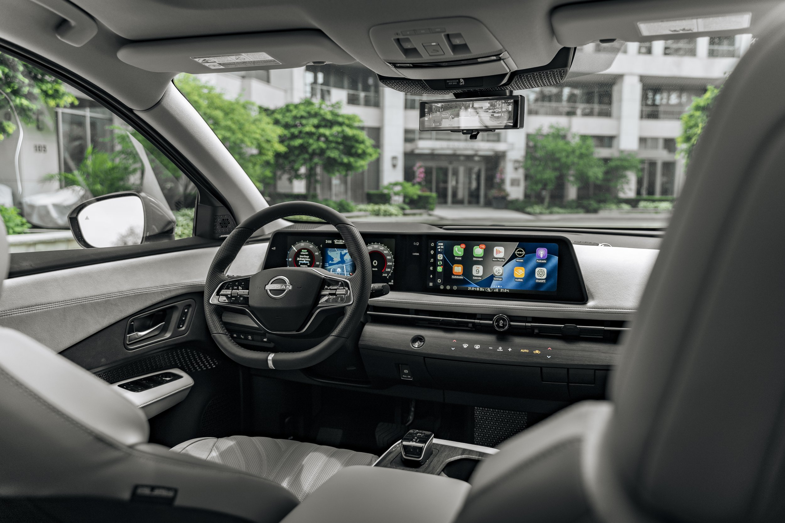 Interior view of a modern Nissan vehicle, showing the dashboard, steering wheel, and an infotainment screen, with a view of a city street with trees and buildings through the windshield.