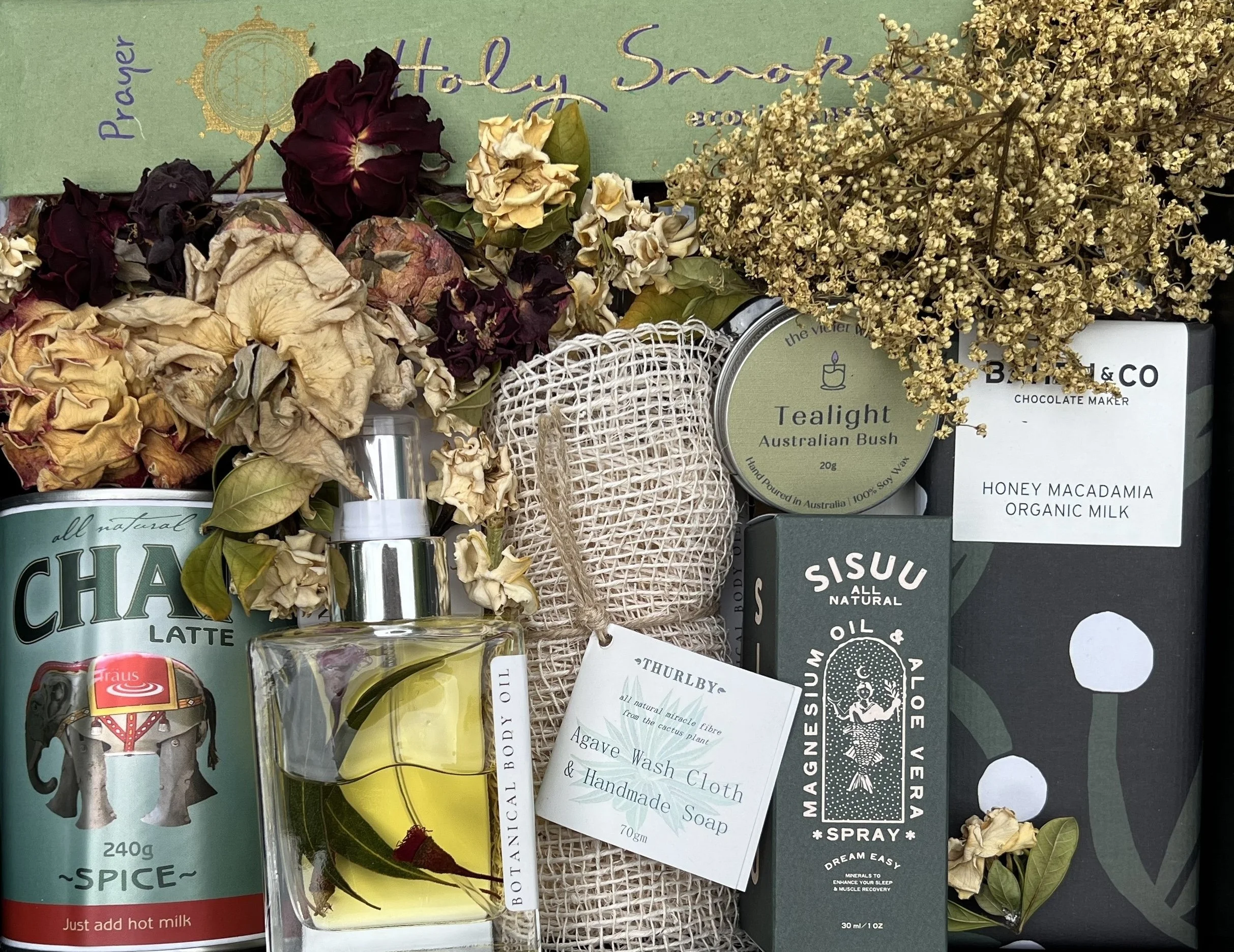 A collection of dried flowers, a bottle of perfume with yellow liquid, a tin of tealaight Australian Bush, a box of honey macadamia organic milk, a box of Sisuu essential oil spray, a box of Thurlby Agave Wash Cloth & Handmade Soap, an all natural chocolate latte can, and a herbal mesh bag with more dried flowers on a surface.