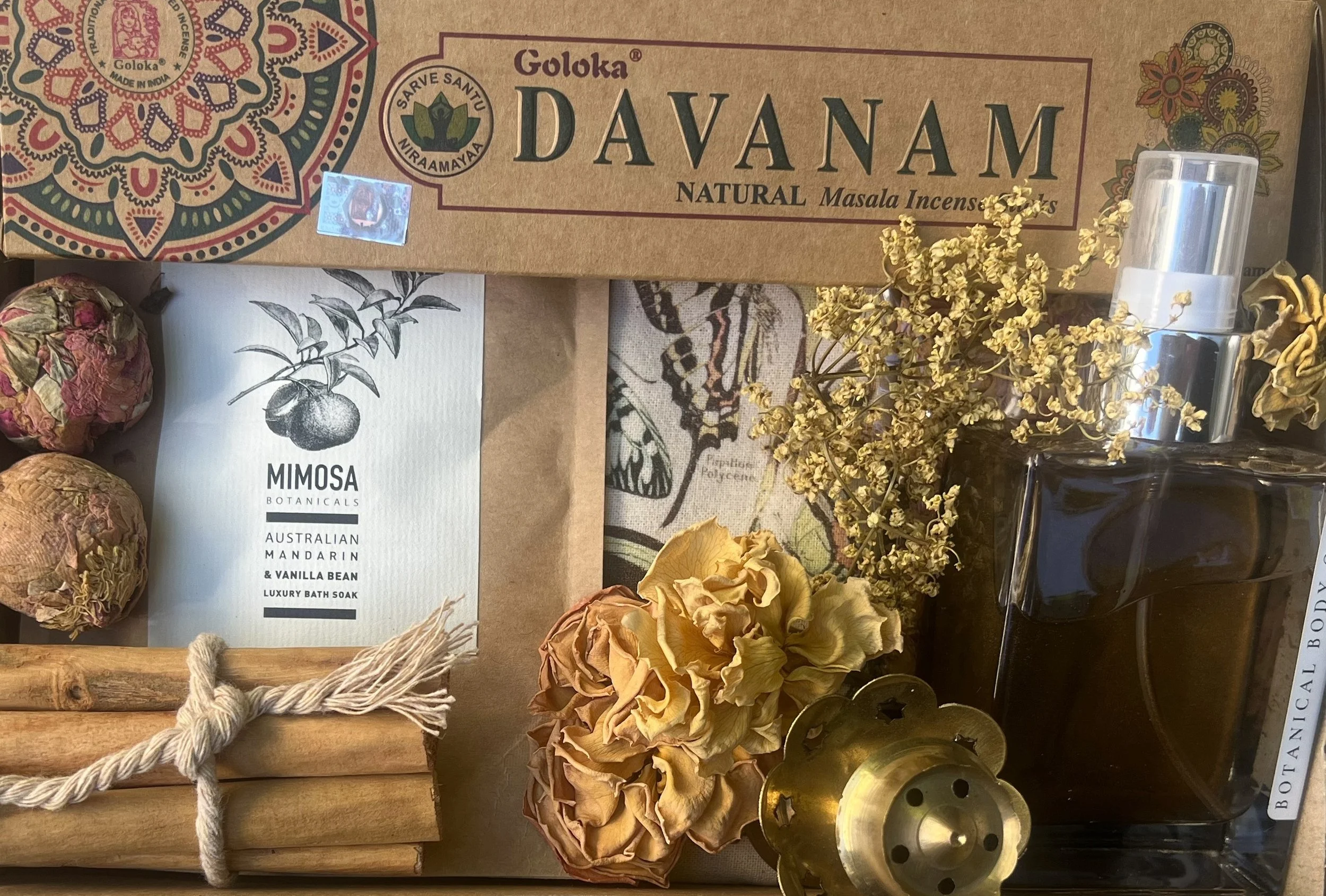 Arrangement of natural and herbal products, including a box of Goloka Davanam incense sticks, a package of Mimosa botanical bath soak, dried flowers, cinnamon sticks, a perfume bottle, and decorative items.