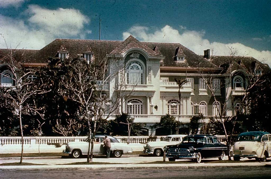 A large Victorian-style mansion with multiple stories, gabled roof, and ornate architecture. Several vintage cars are parked in front, with a man standing near one of them. Leafless trees line the street, and the sky is partly cloudy.
