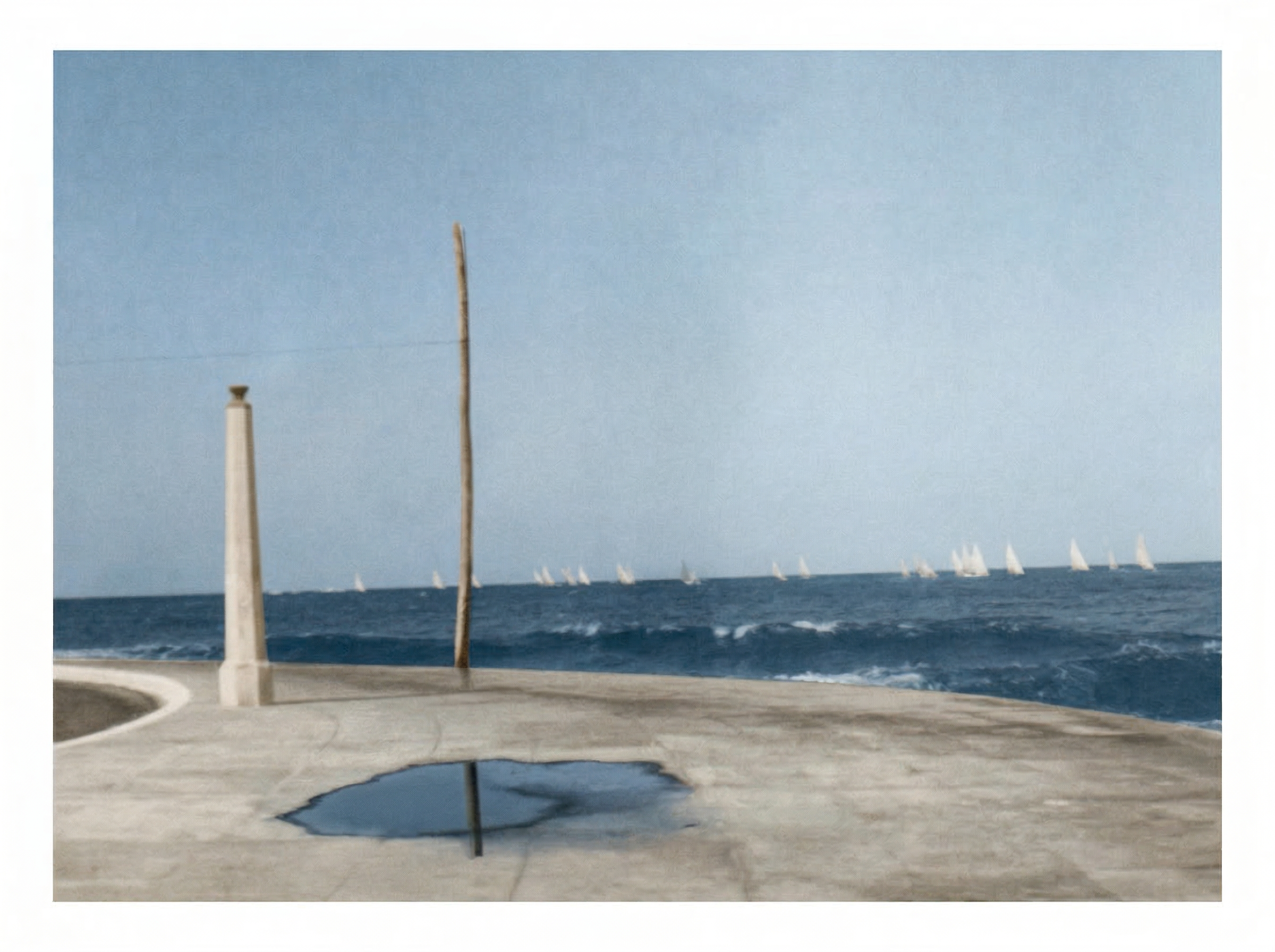 A view of a seascape with sailboats on the horizon, seen from a concrete waterfront with a puddle reflecting a wooden pole and a short concrete column.