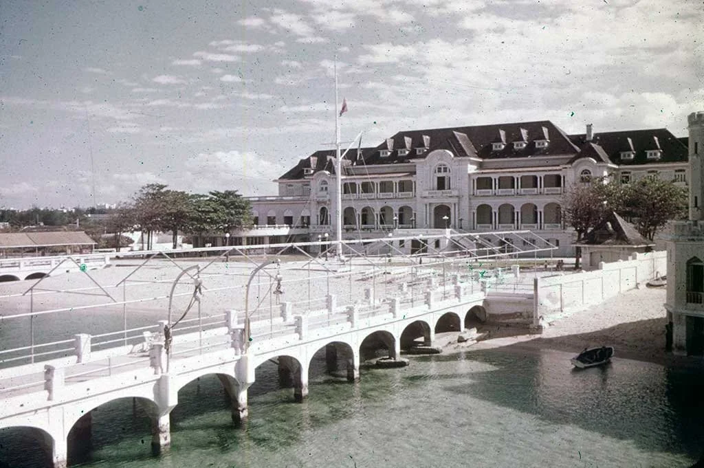 A historic hotel with a large, ornate building, a bridge over water, and a boat docked nearby, with some trees and a partly cloudy sky.