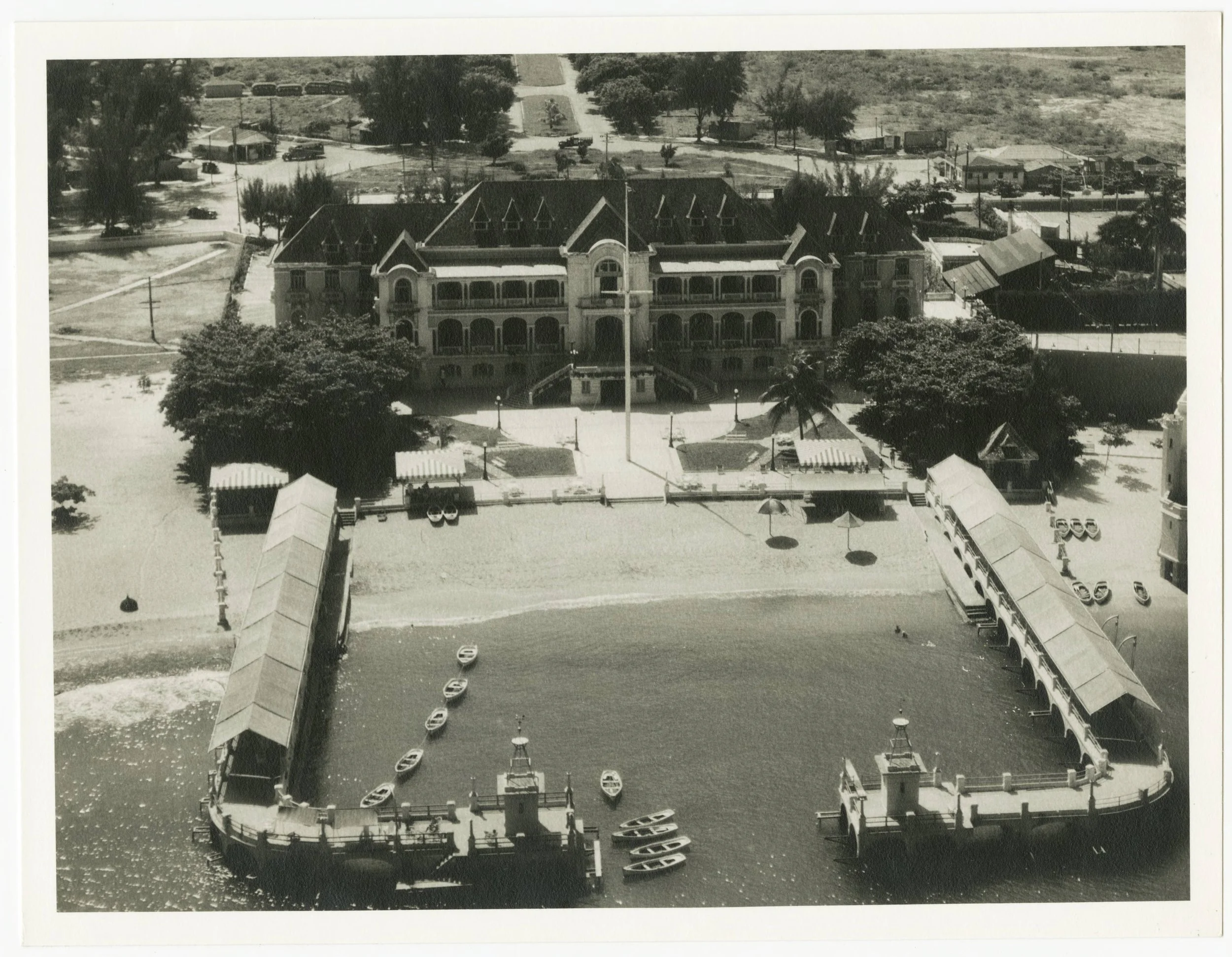 Aerial view of a large Victorian-style building with a dock, boats, and a sandy area in front, surrounded by trees and smaller structures.