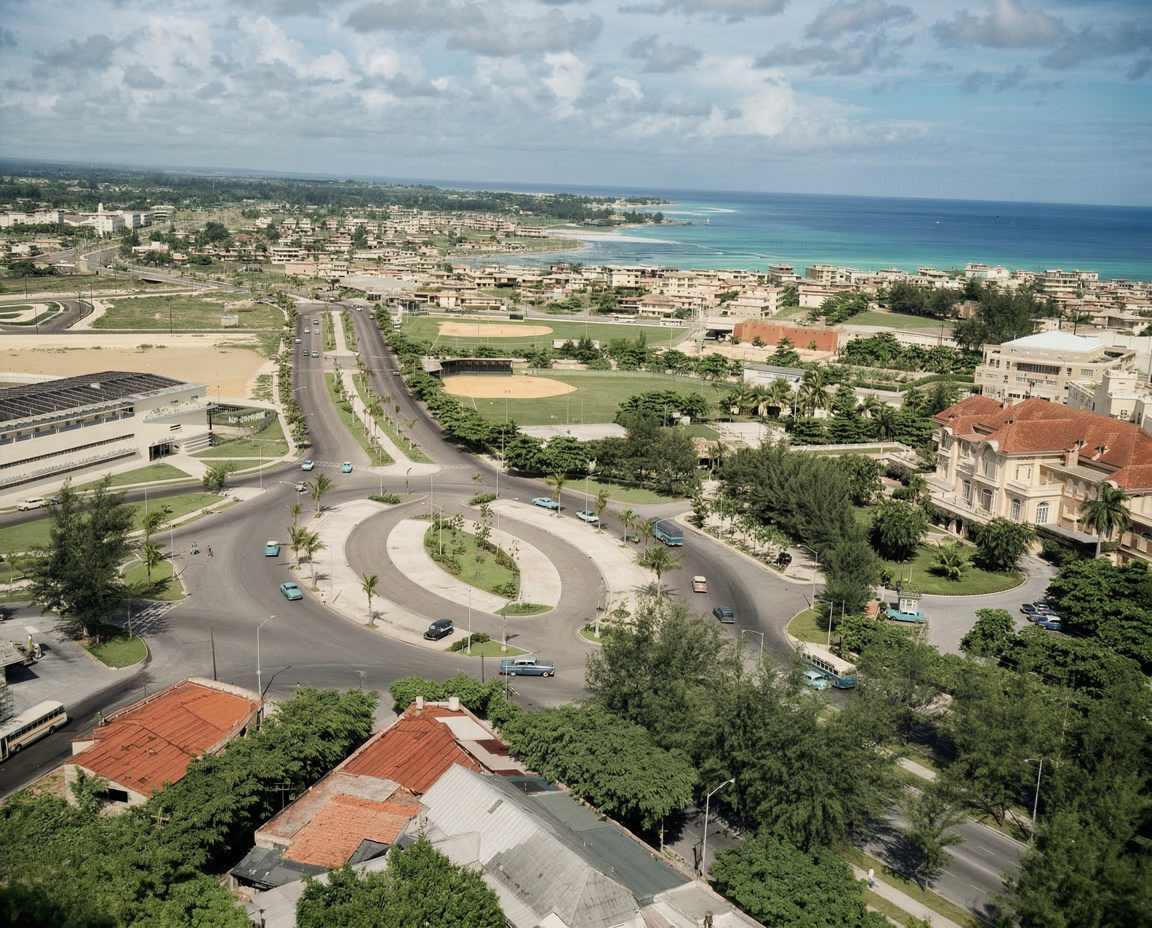 An aerial view of a coastal city with a roundabout, roads, residential buildings with red roofs, a golf course, and the ocean in the background under a partly cloudy sky.