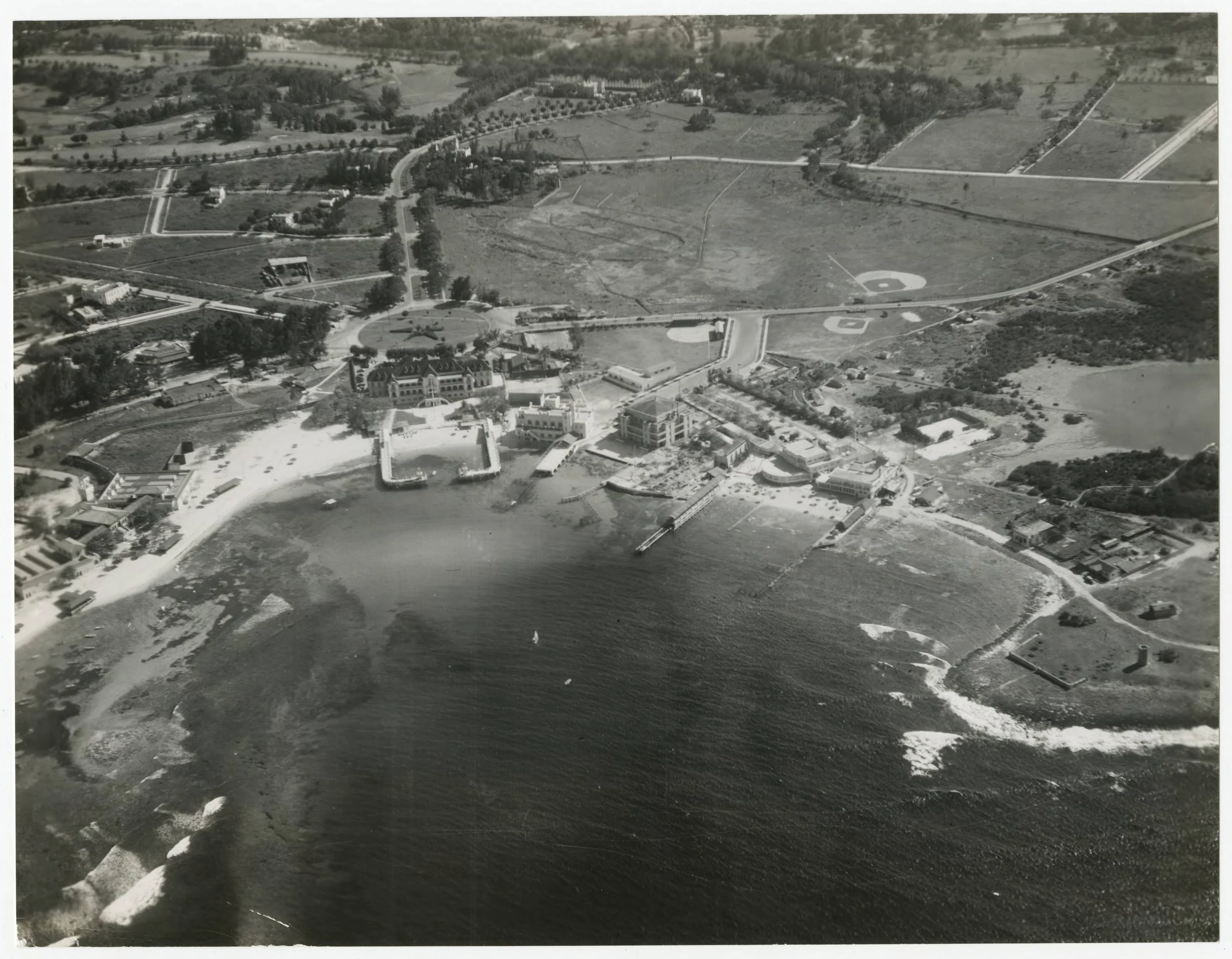 Aerial black and white photograph of a coastal area showing a large hotel near the water, with piers extending into the ocean, a beach, and surrounding roads, buildings, and baseball fields in the background.