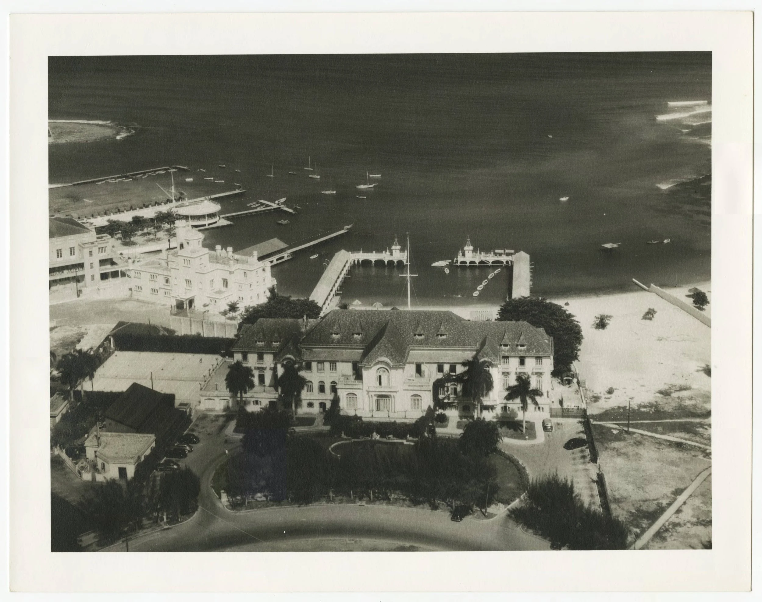 Black and white aerial photograph of a building with a waterfront and marina in the background, featuring boats docked and a pier with arches.