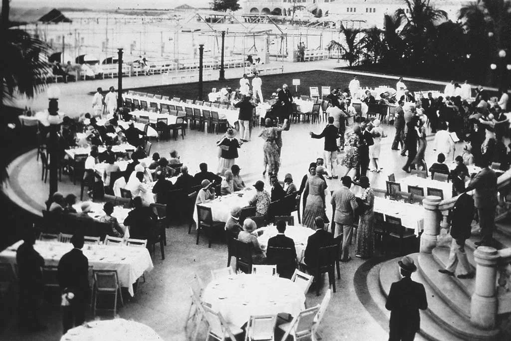 A large outdoor gathering with many people dancing and socializing around tables set for dining at an elegant event, with a pool and tropical trees in the background.
