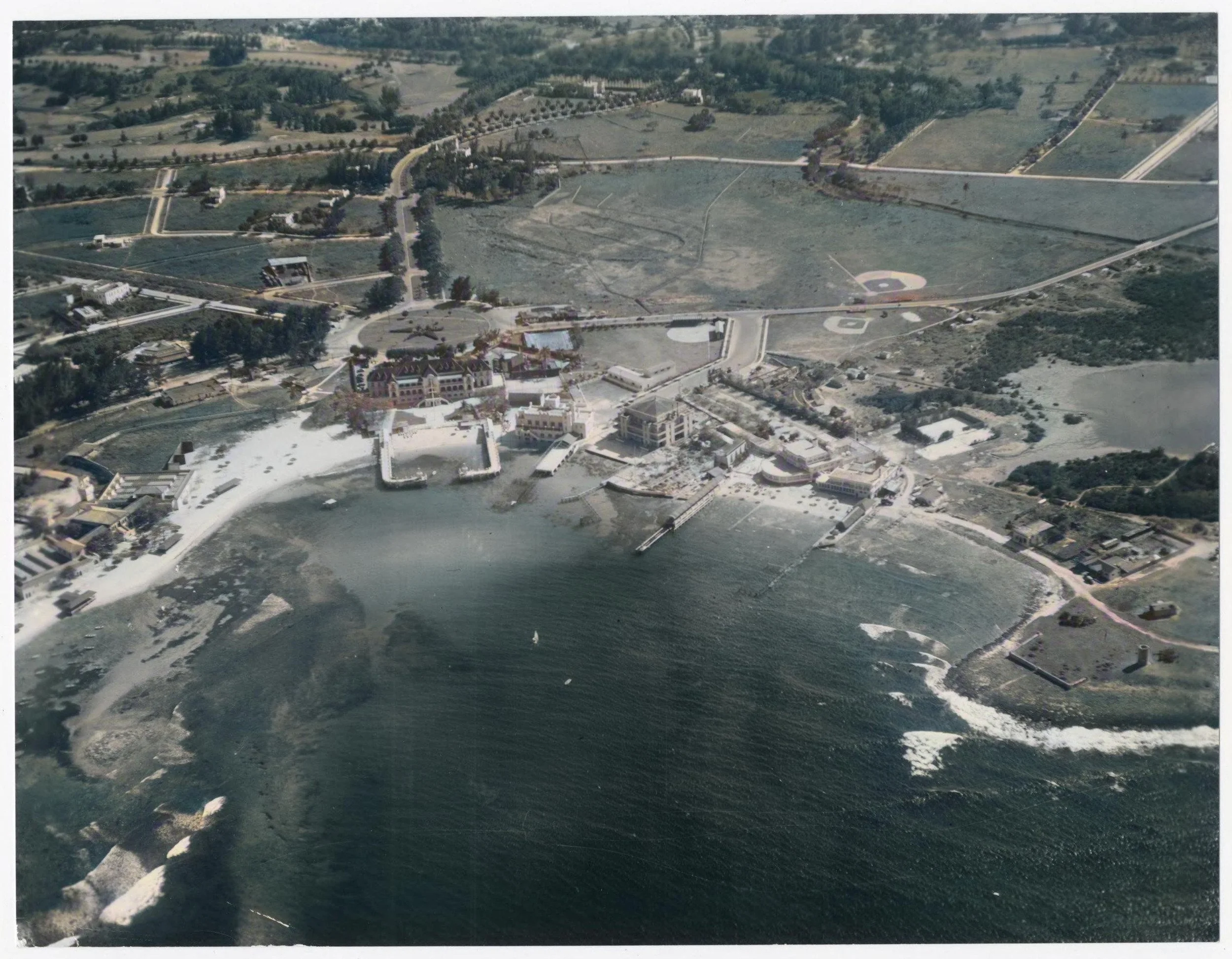 Aerial view of a large coastal resort with a harbor, multiple buildings, a river, and baseball fields surrounded by greenery and roads.