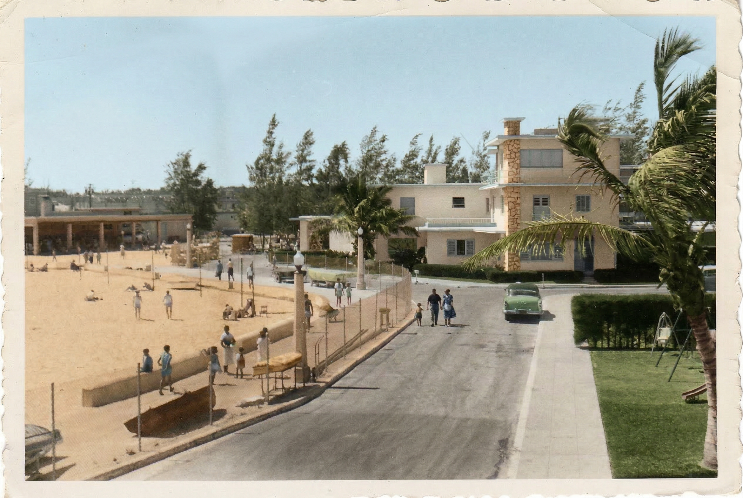 A vintage color photograph of a beachside neighborhood street with people walking and lounging on the sandy beach. There are palm trees, a grassy area with swings, a large modern house, and vintage cars parked along the street.