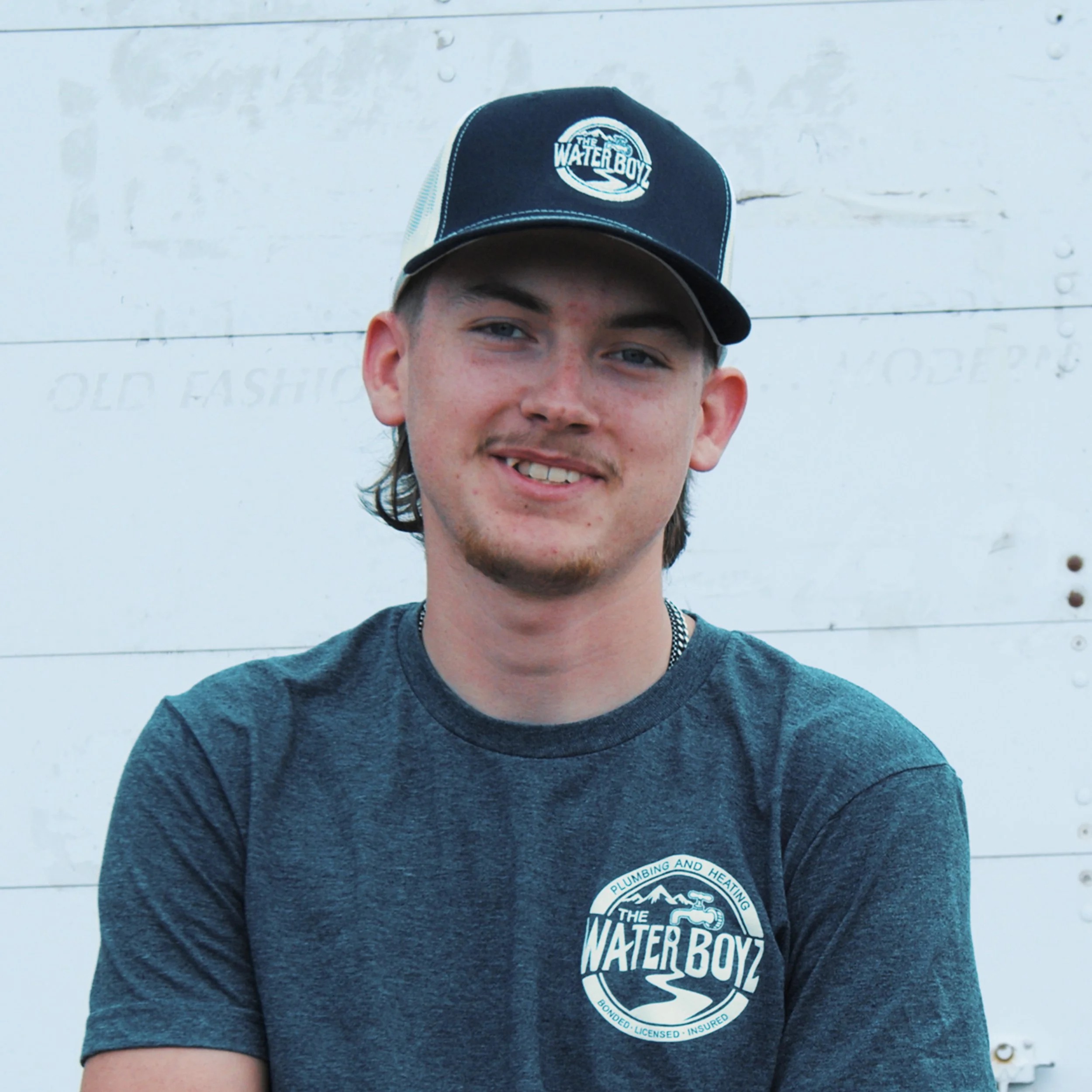 A young man wearing a black and white baseball cap with a logo that reads 'The Water Boyz' and a dark blue T-shirt with a circular logo for plumbing, heating, and cooling services, standing in front of a white wooden wall.