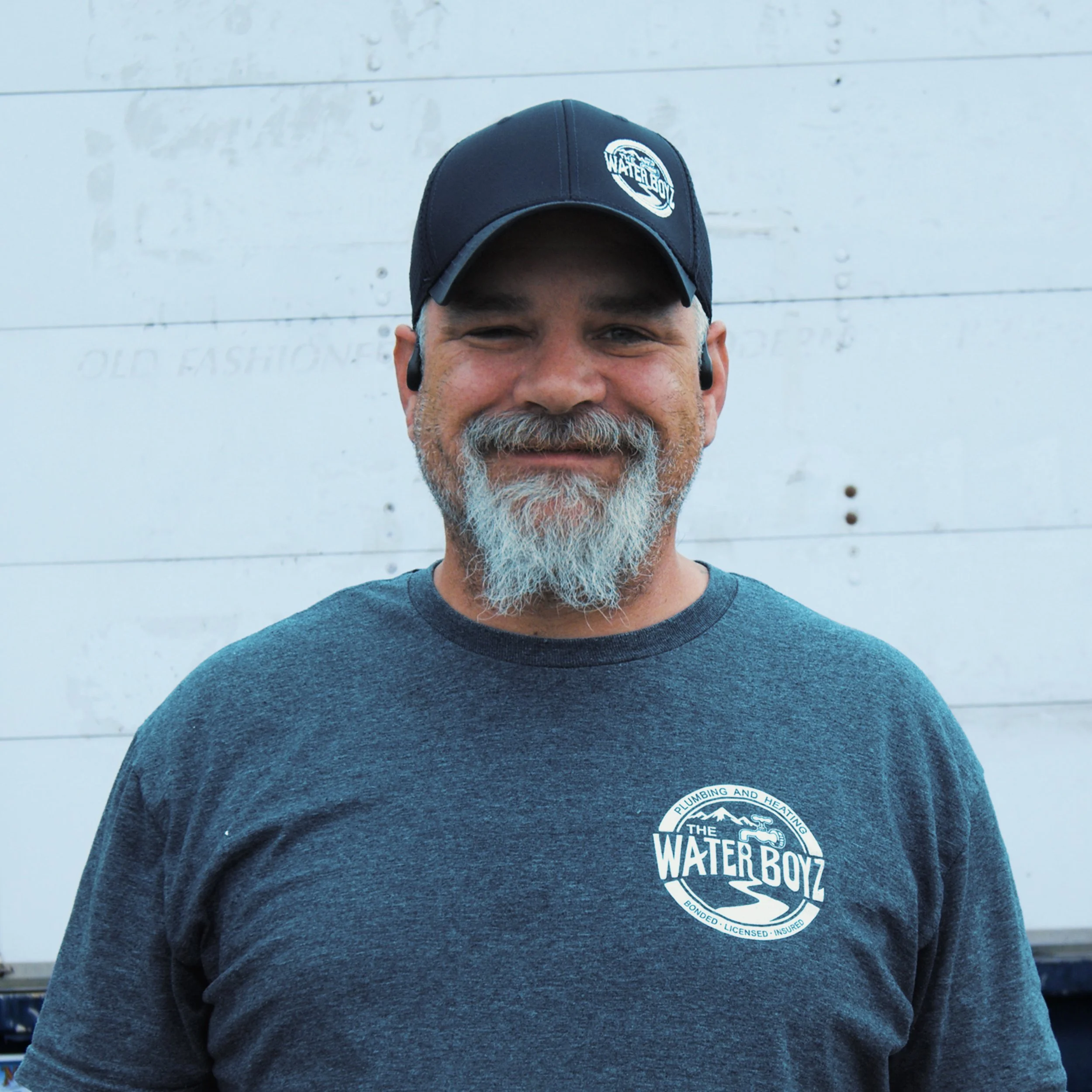 Close-up of a smiling man wearing a dark baseball cap with the logo 'Water Boyz' and a dark t-shirt with the same logo, standing against a white wooden background.