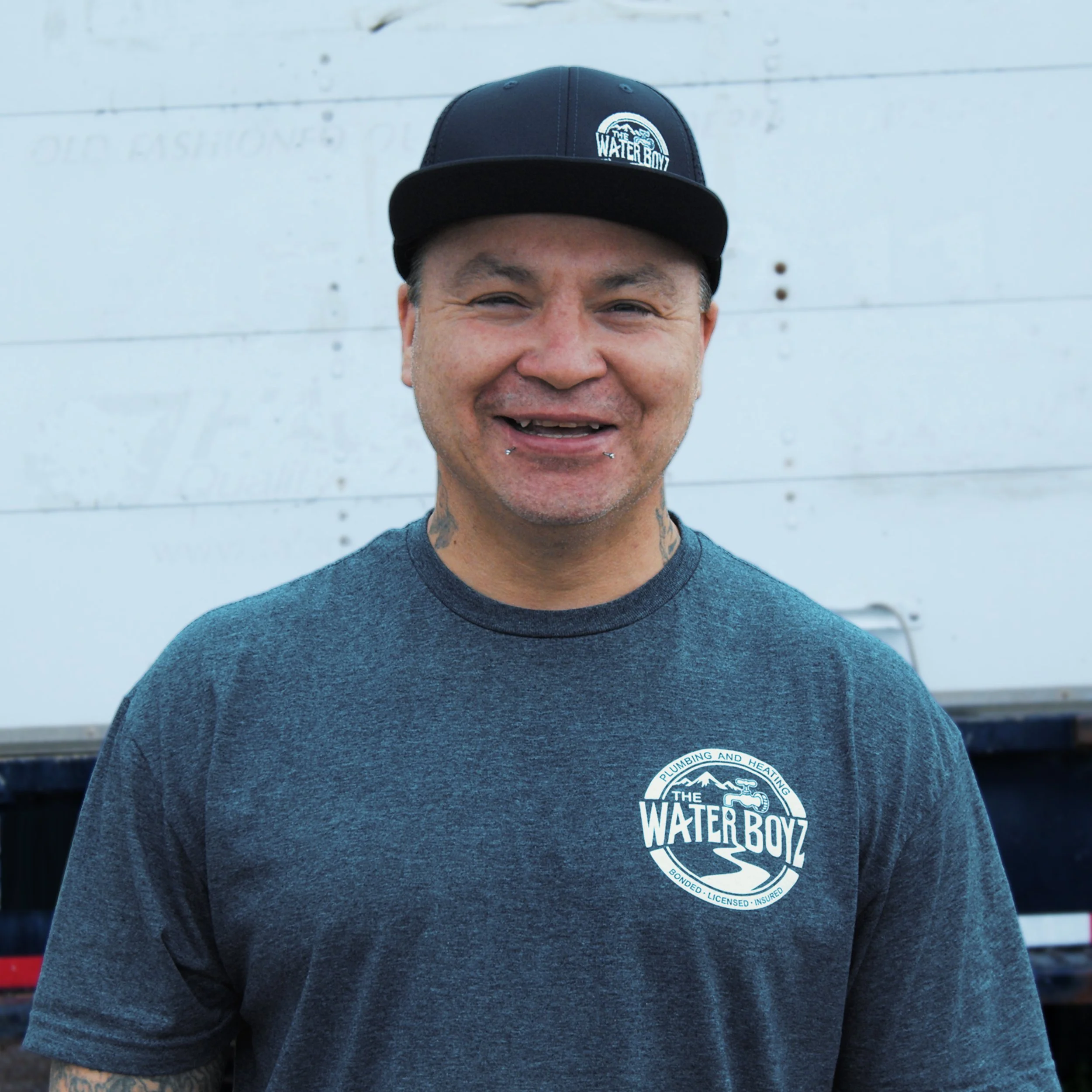 A smiling man wearing a black baseball cap with a logo that reads 'Water Boyz' and a dark gray T-shirt with the same logo, standing in front of a white wall.
