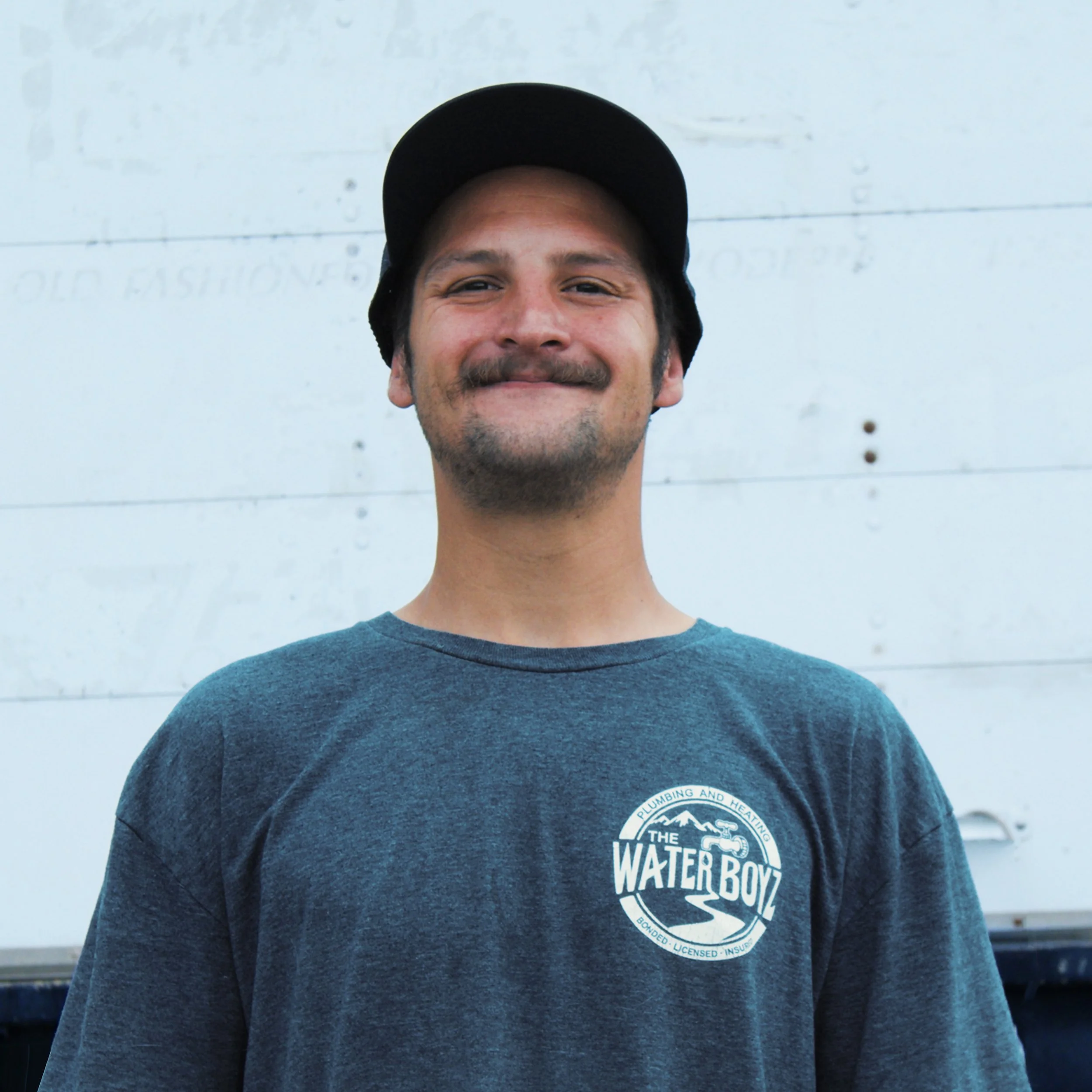 Close-up of a smiling man wearing a dark gray t-shirt with a circular logo reading 'The Water Boy' and a black baseball cap, standing in front of a light-colored wall with faint text.